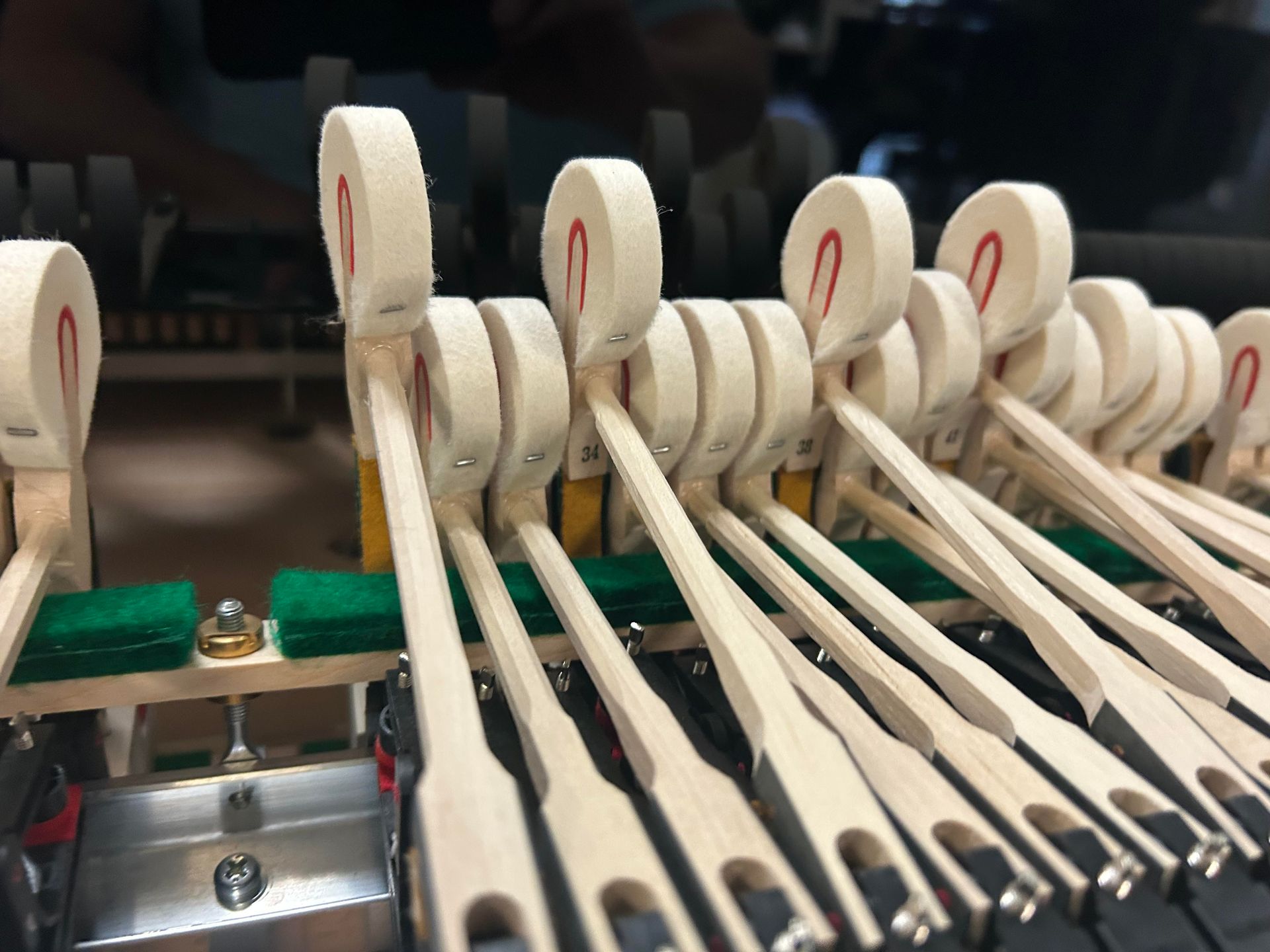 Piano hammers with red felt striking strings, wooden levers, and green felt supports.