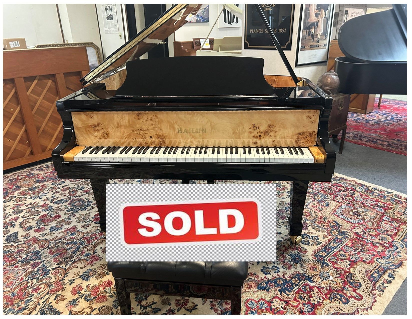A grand piano with a black lid and a light wood front sits on a floral rug. A black leather piano stool is in front of the piano.