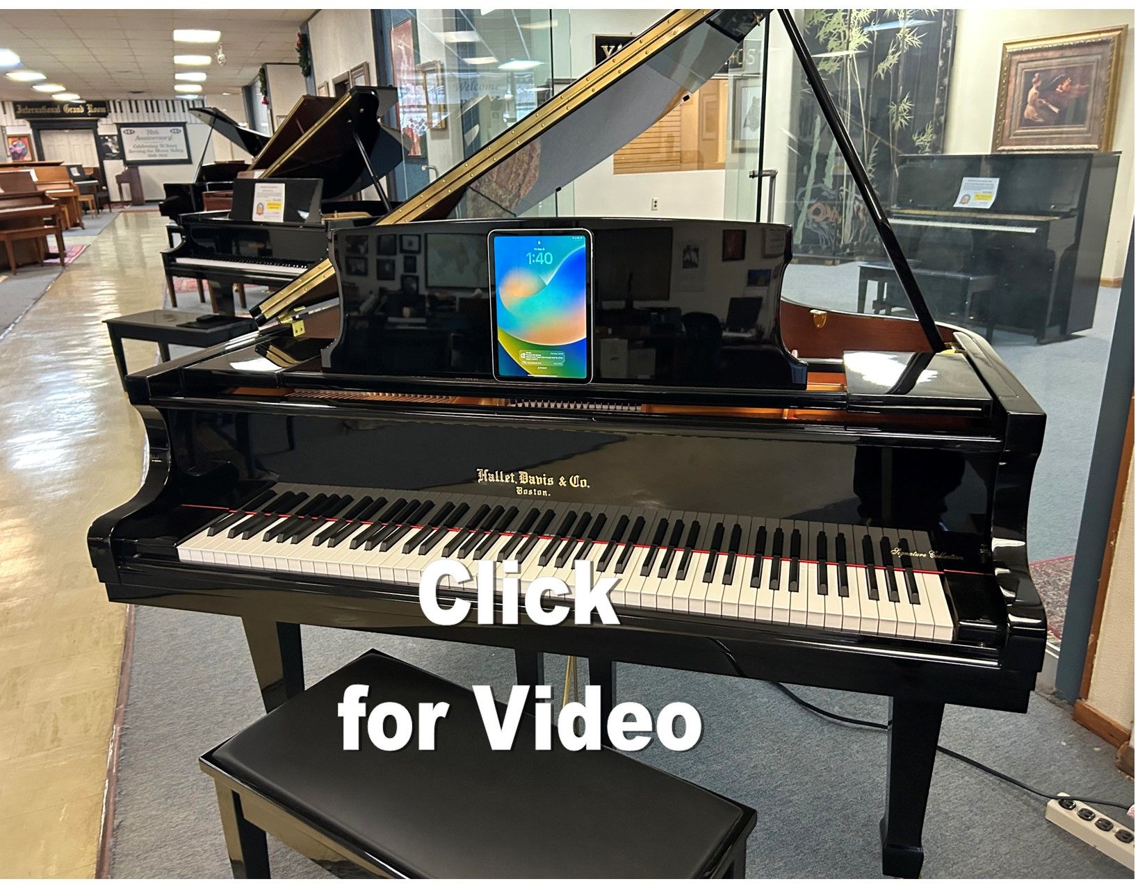 Black grand piano in a showroom, with a tablet on the music rest and a black bench in front.