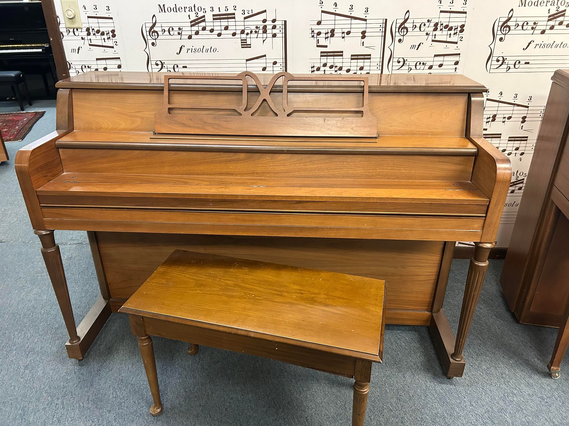 A wooden piano with a bench in front of a wall of sheet music.
