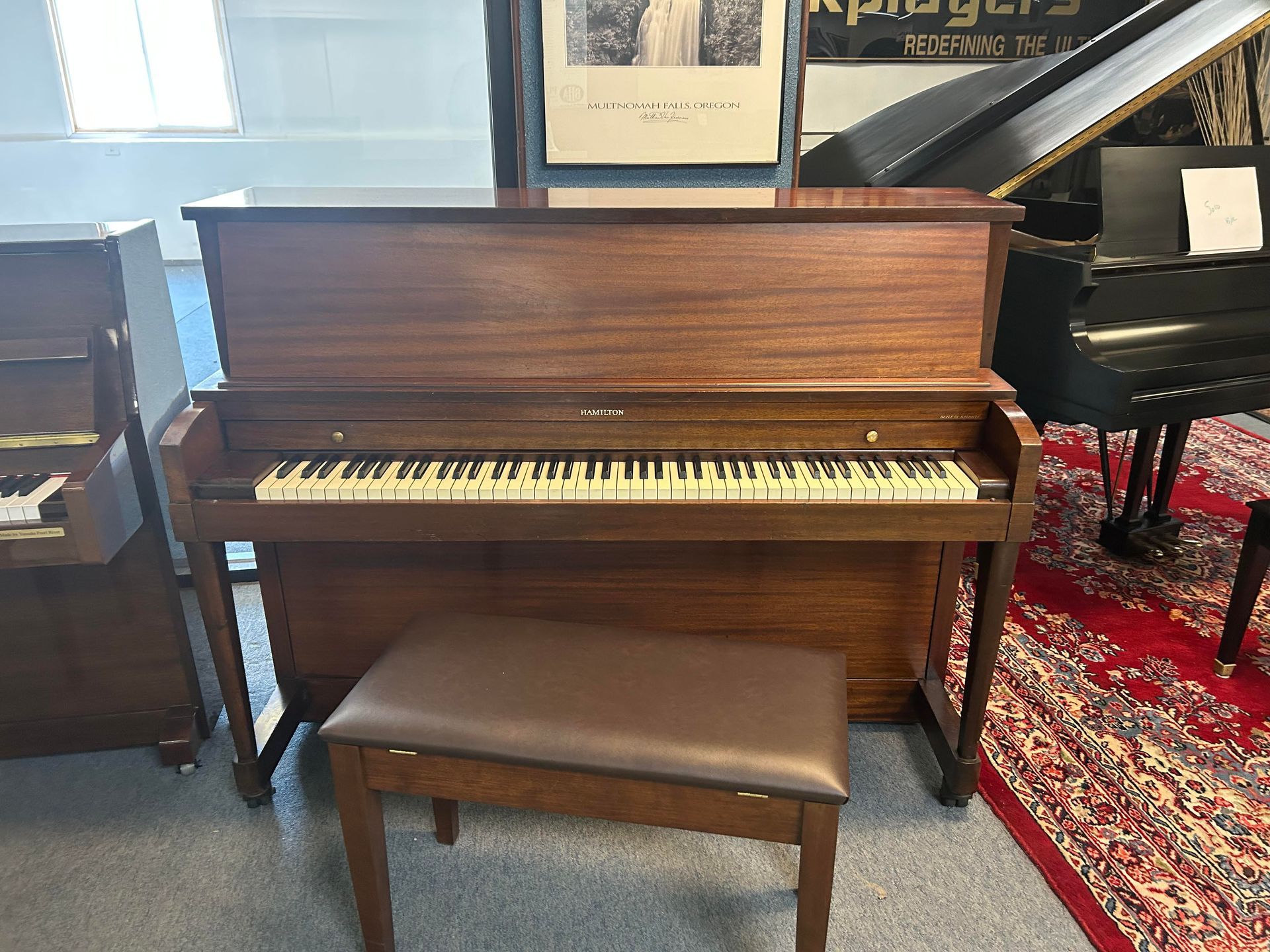 Brown upright piano with matching bench.  Piano has open keyboard and is in a room with other pianos.