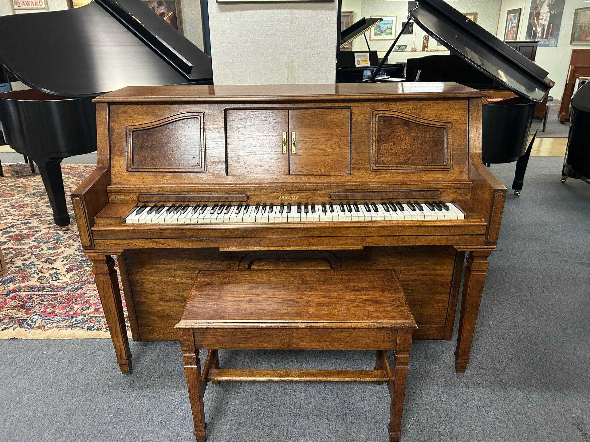Wooden upright piano with bench in a room with other pianos.
