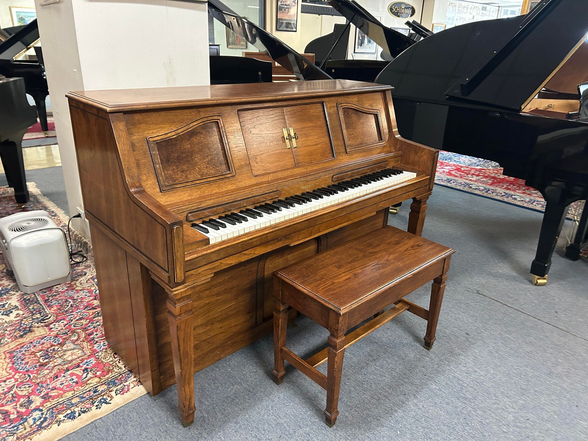 Wooden upright piano and bench in a showroom, with a black grand piano in the background.