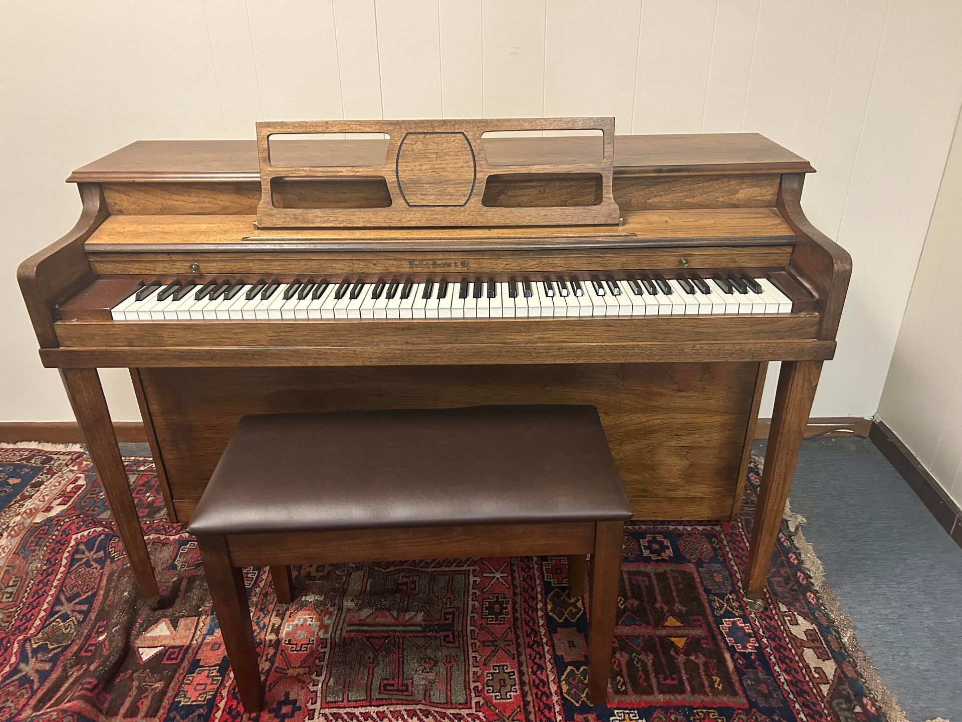 Wooden piano with bench on a patterned rug, against a light wall.