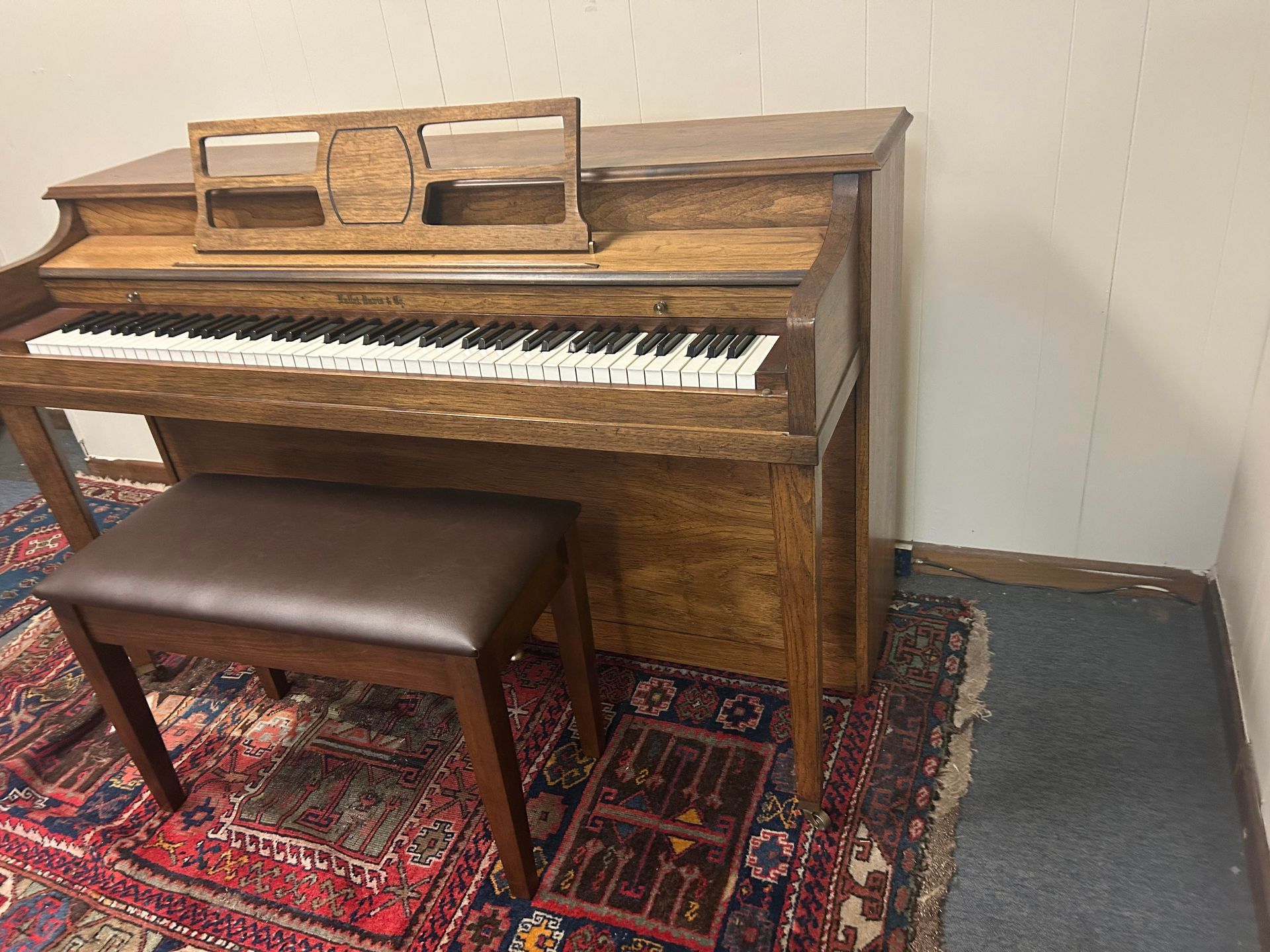 Wooden upright piano with music stand, stool, and rug in a corner.