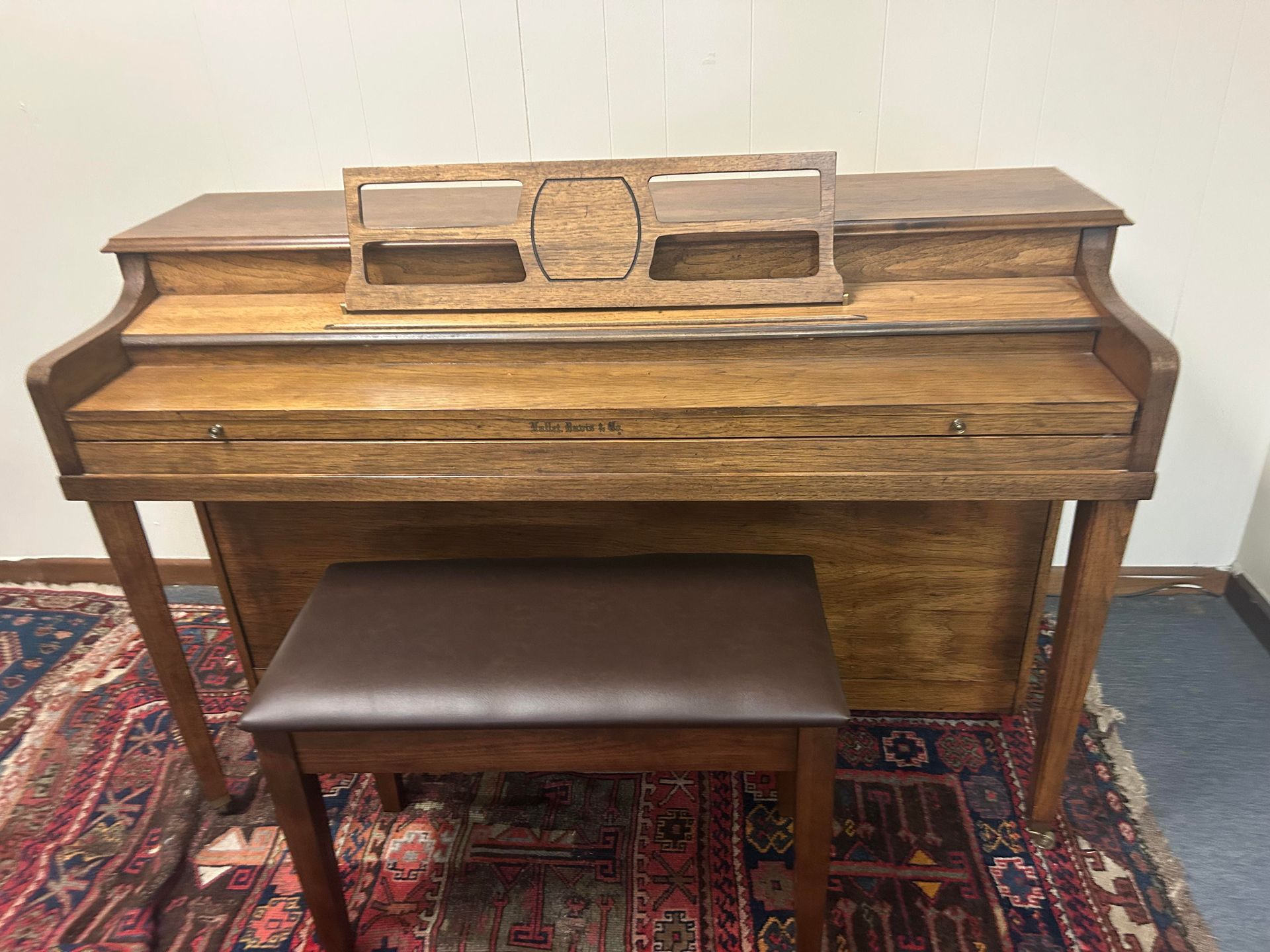 Wooden upright piano with music stand and brown seat, on a patterned rug.