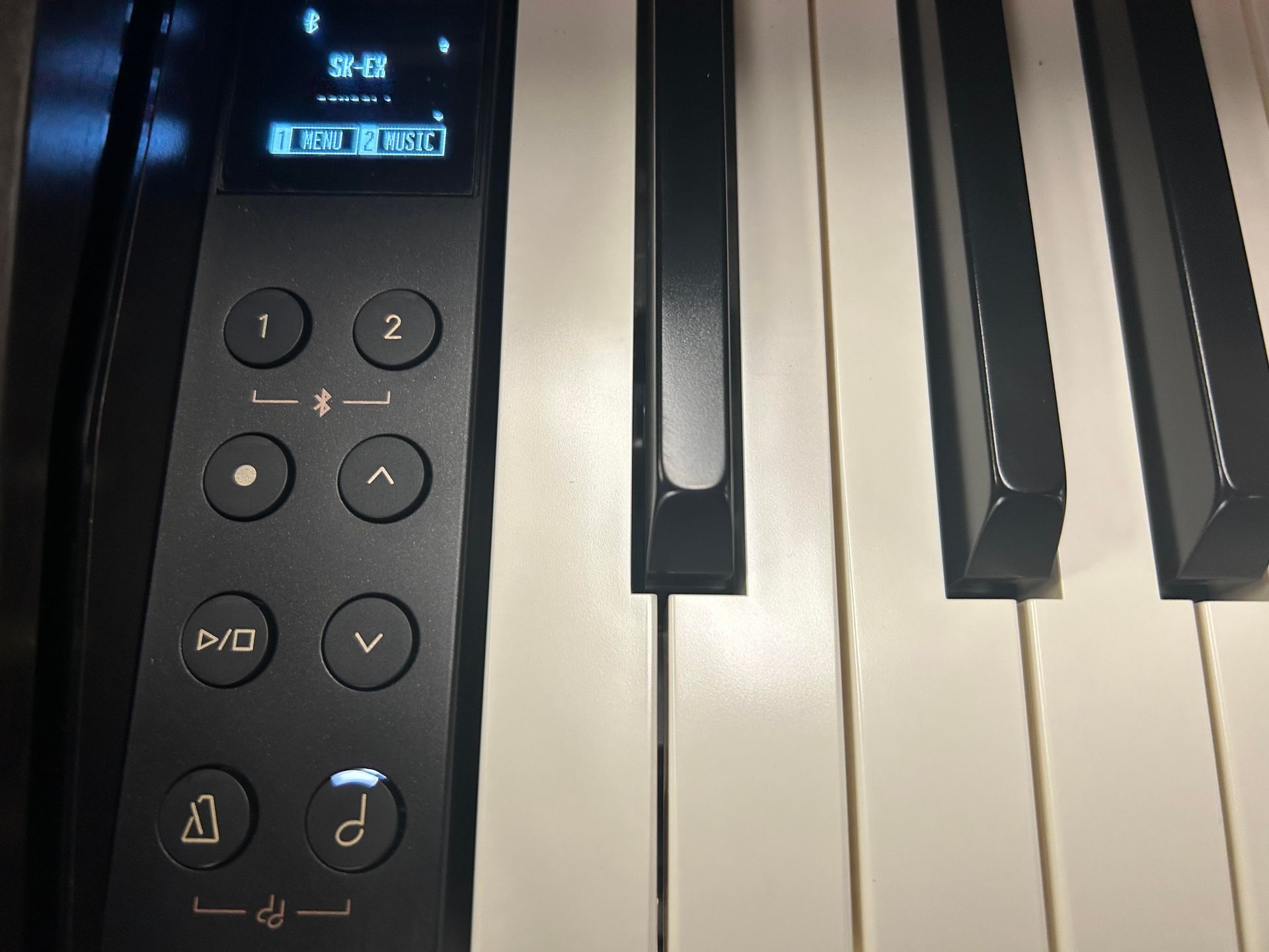 Close-up of a digital piano's control panel and keys. The panel is black, with buttons and a small display. The keys are black and white.