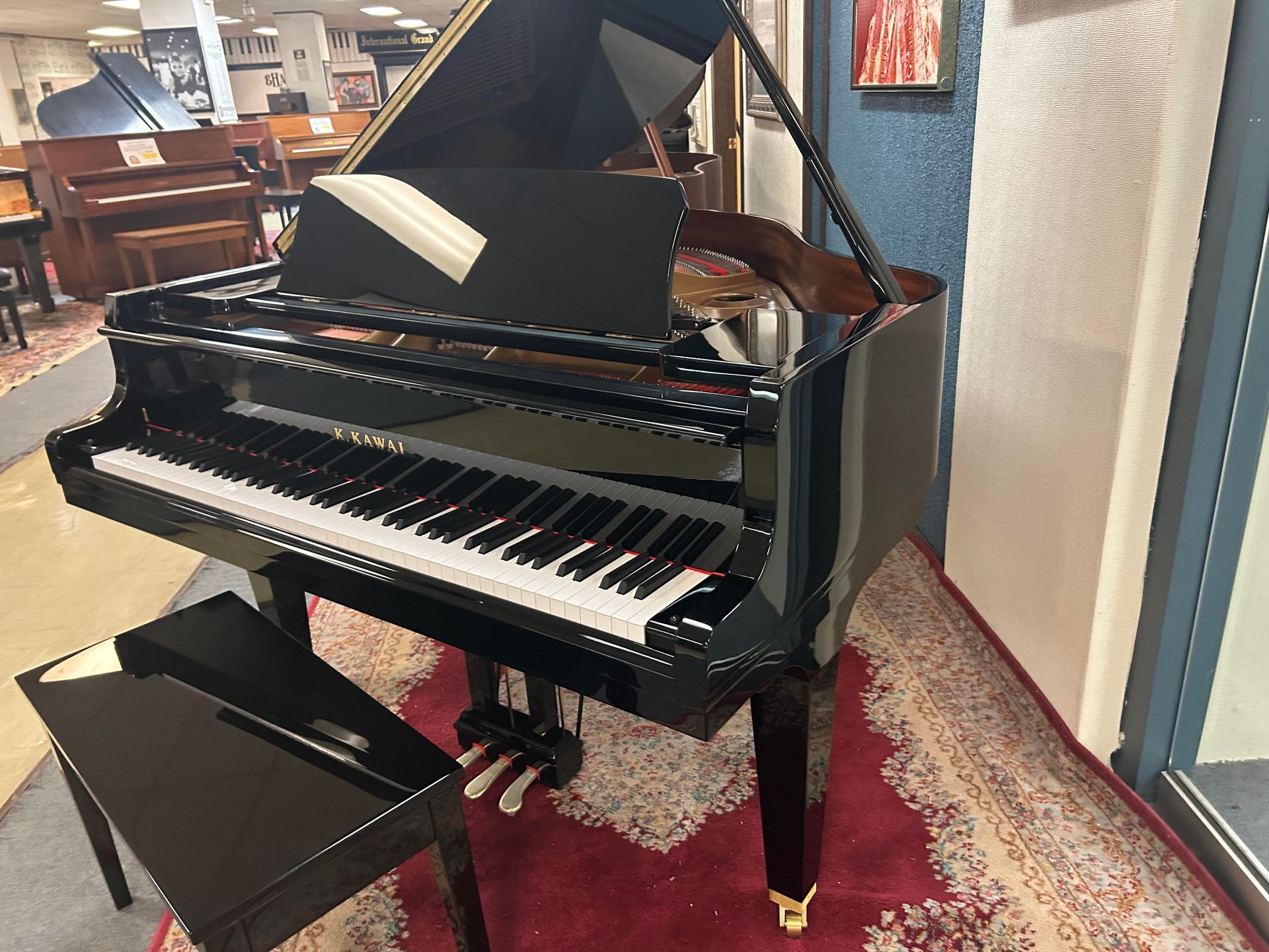 Black grand piano with open lid in a showroom, next to a bench, on a rug.