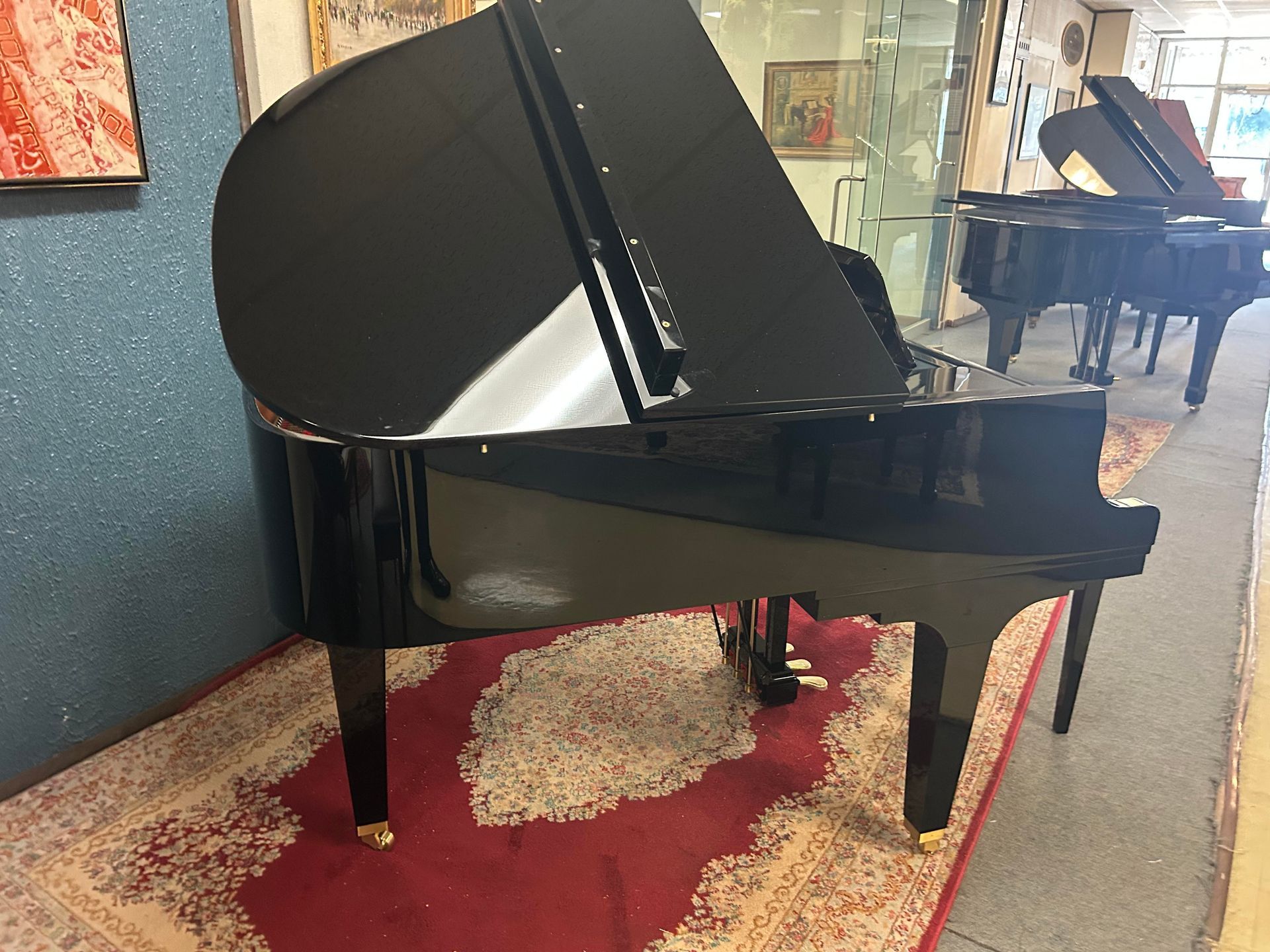 Black grand piano on a red rug in a store, another piano is visible in the background.