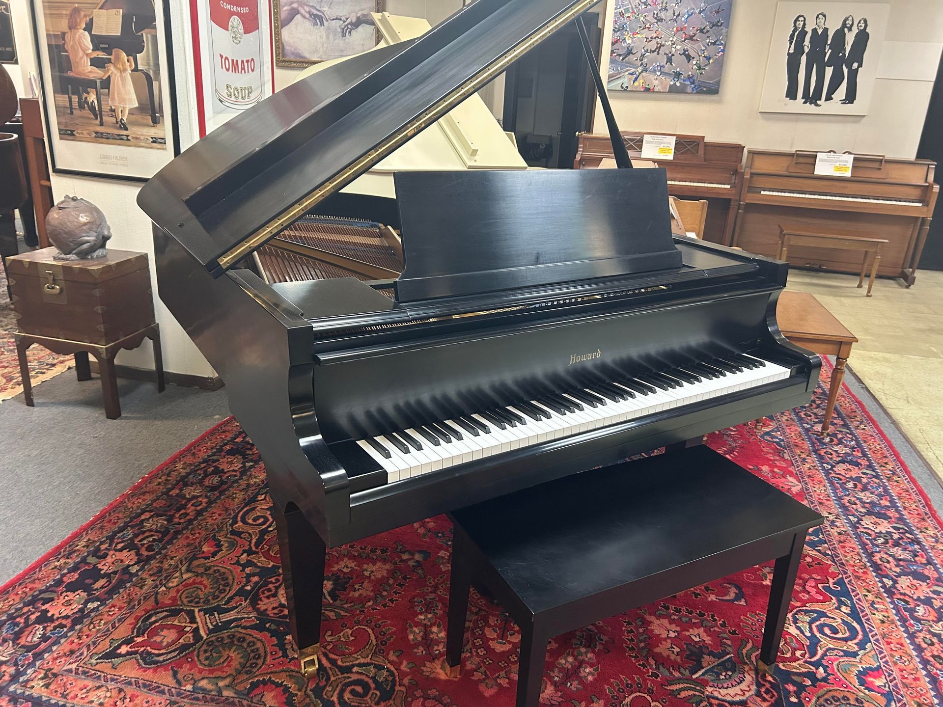 Black grand piano with open lid, black bench, on a red patterned rug. Located in a showroom with other pianos.
