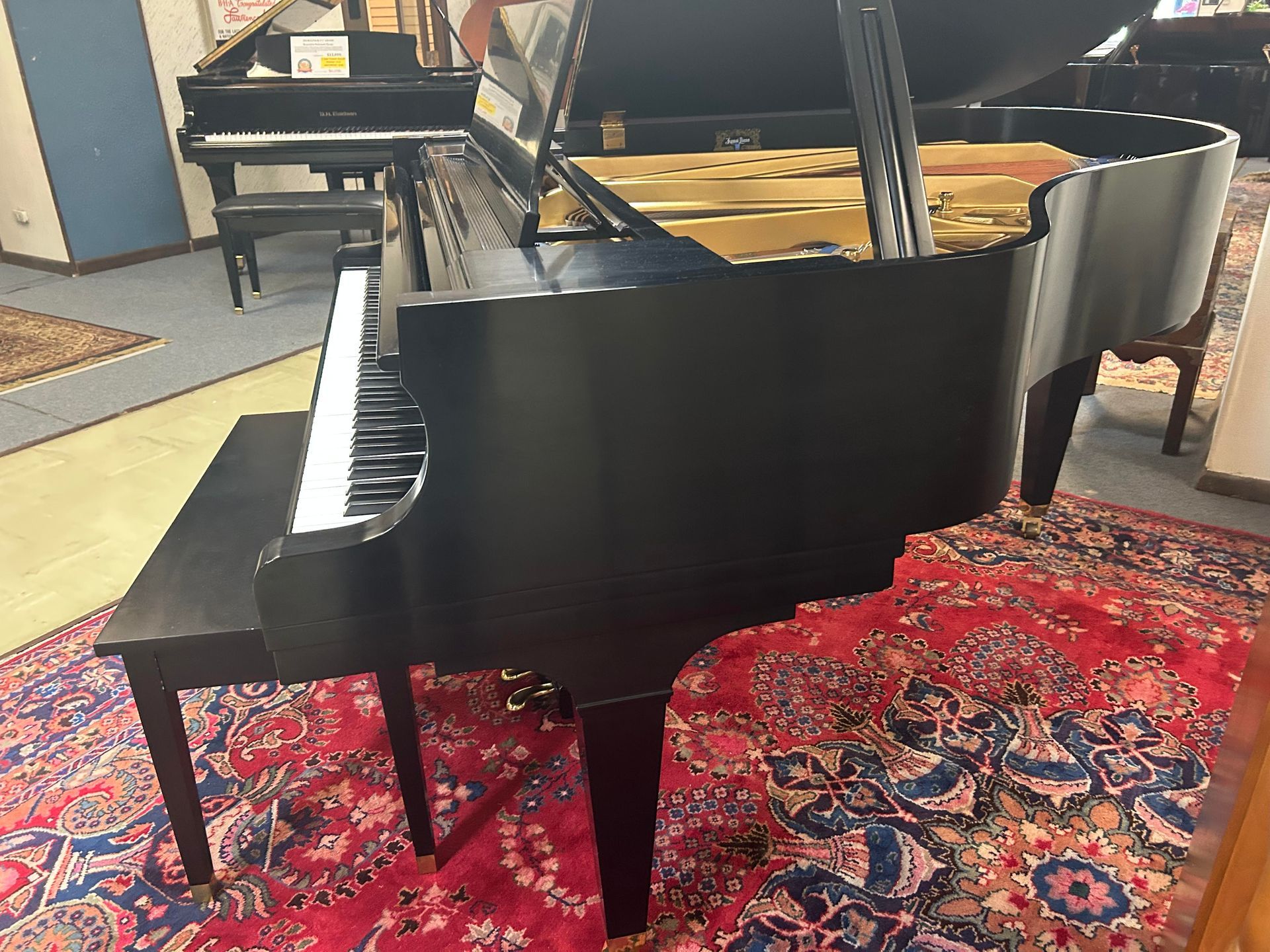 Black grand piano with the lid open, on a patterned red rug, with a smaller piano in the background.