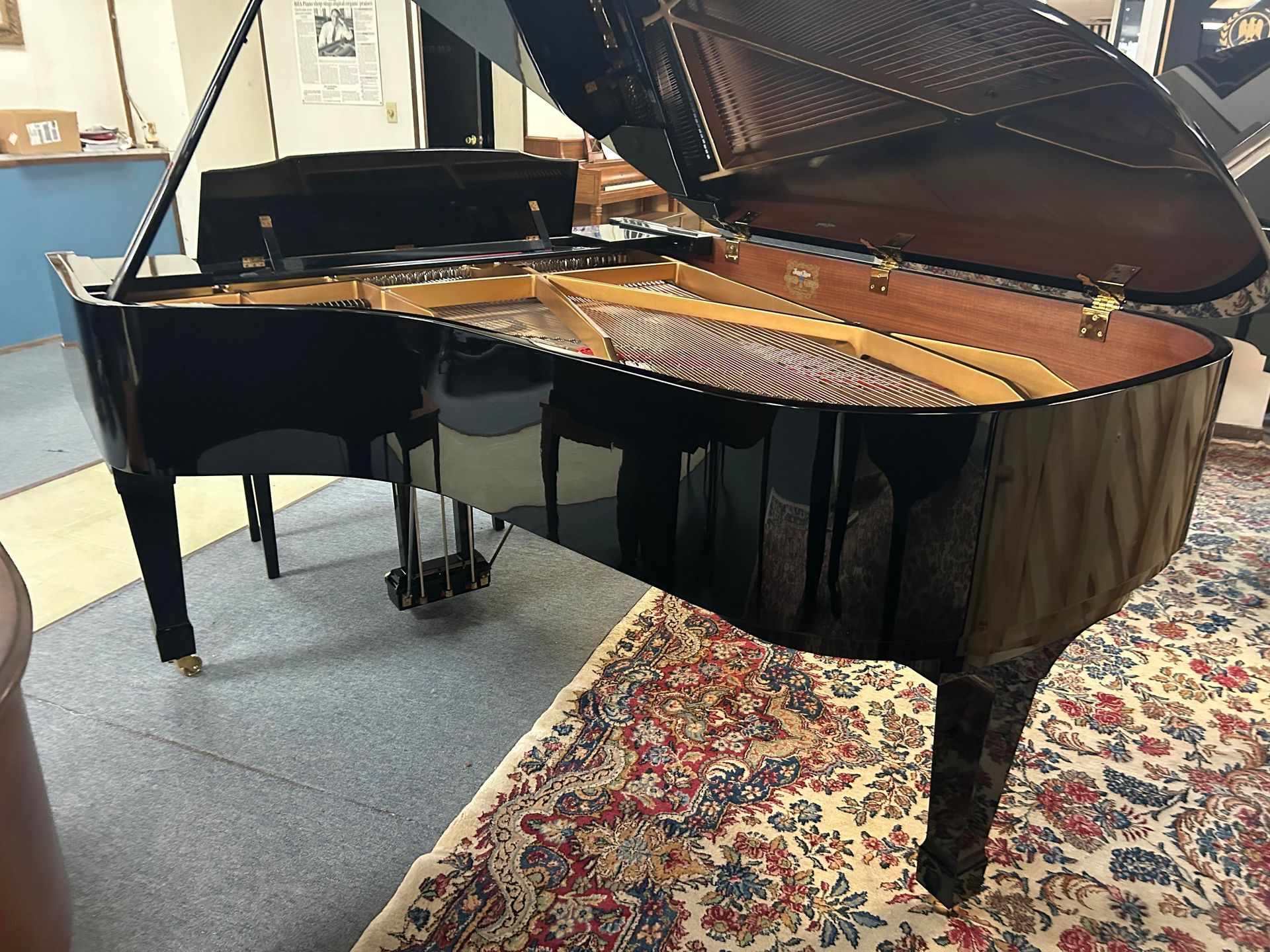 Black grand piano, open, in a room. Shiny surface, visible strings, and a patterned rug in front.