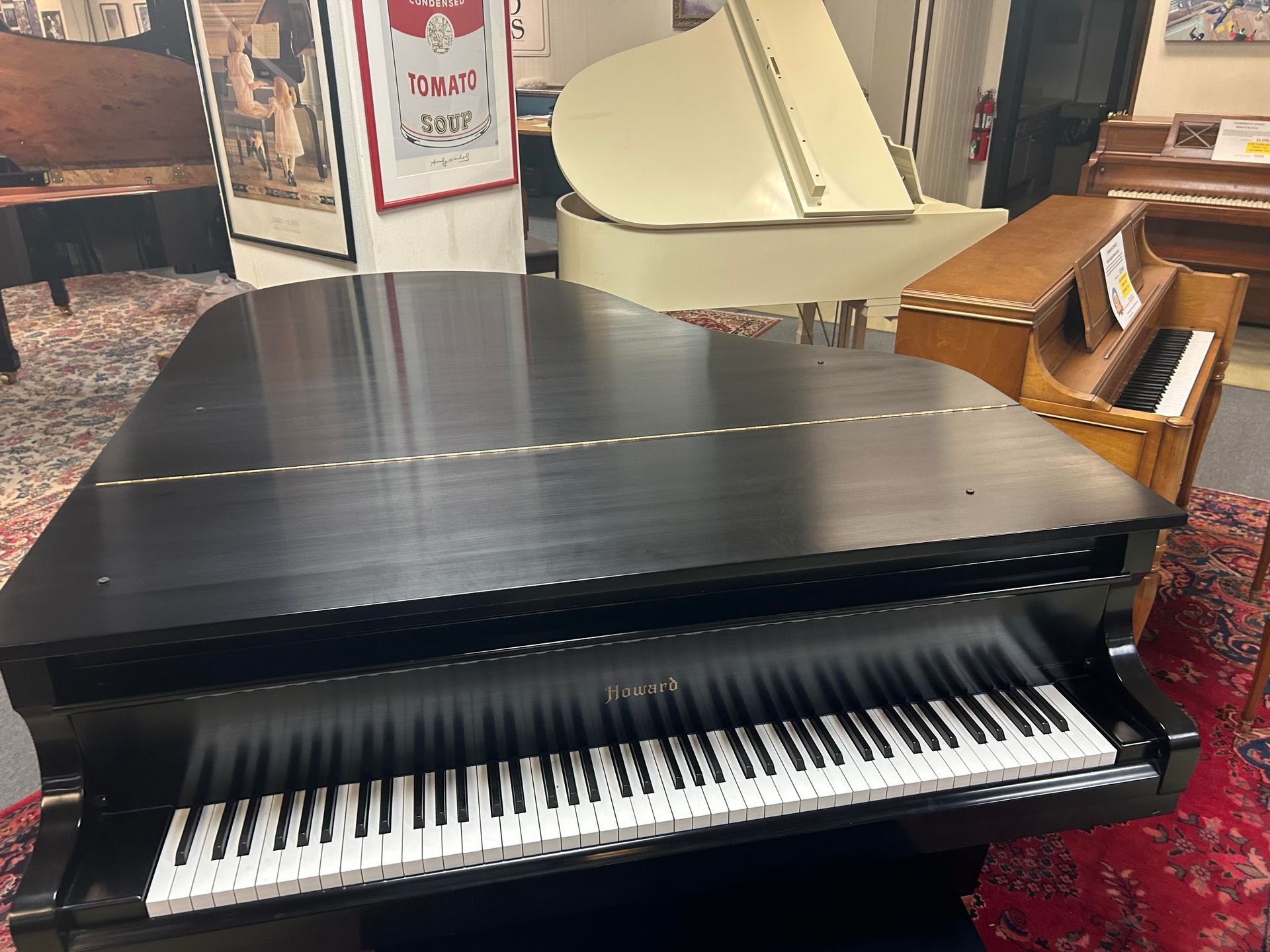 A black grand piano with open lid sits in a shop, with a beige grand and a smaller upright piano visible in the background.