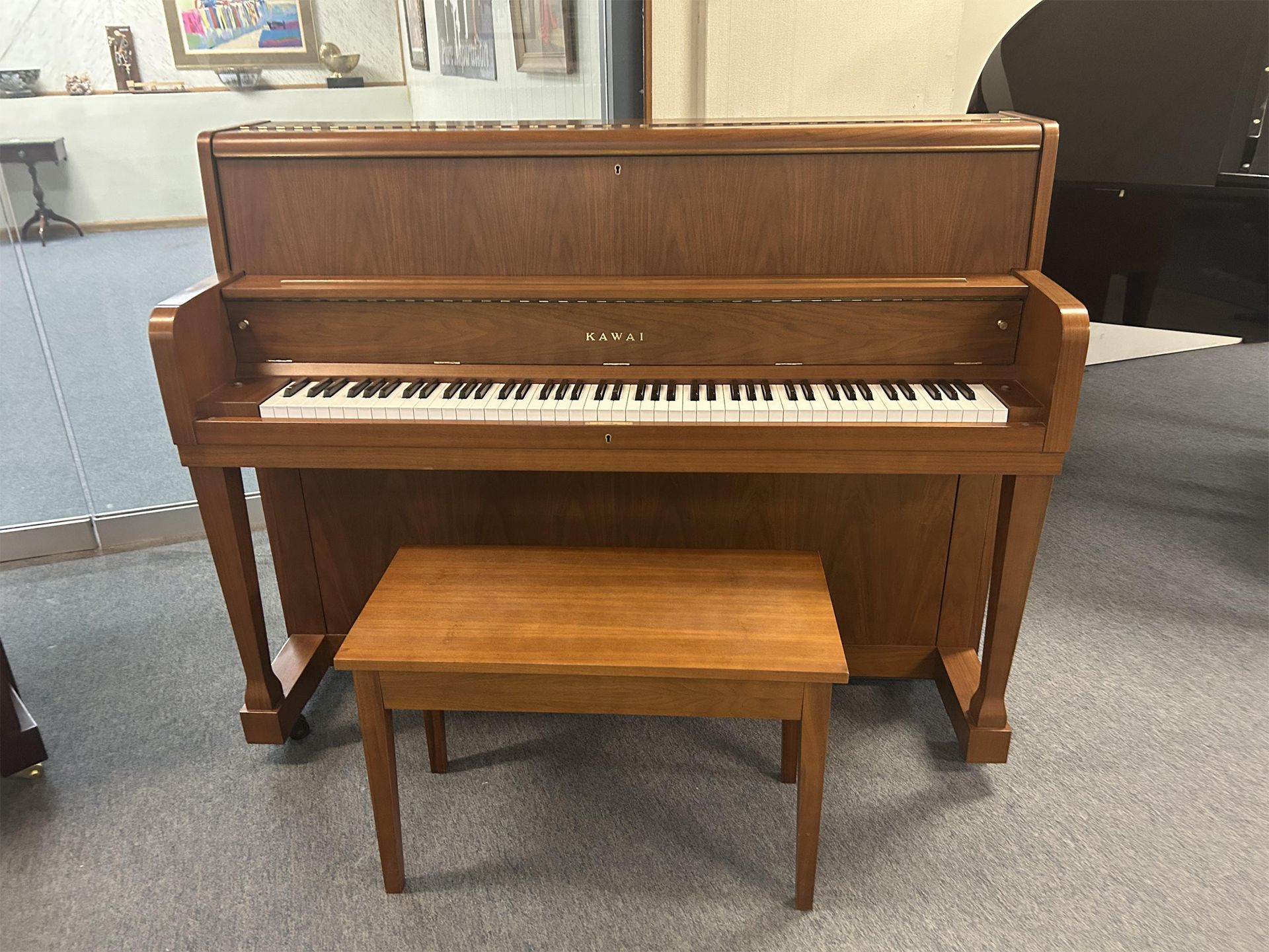 A brown Kawai upright piano with a matching bench sits on a gray carpet in a room decorated with framed art and musical instruments.
