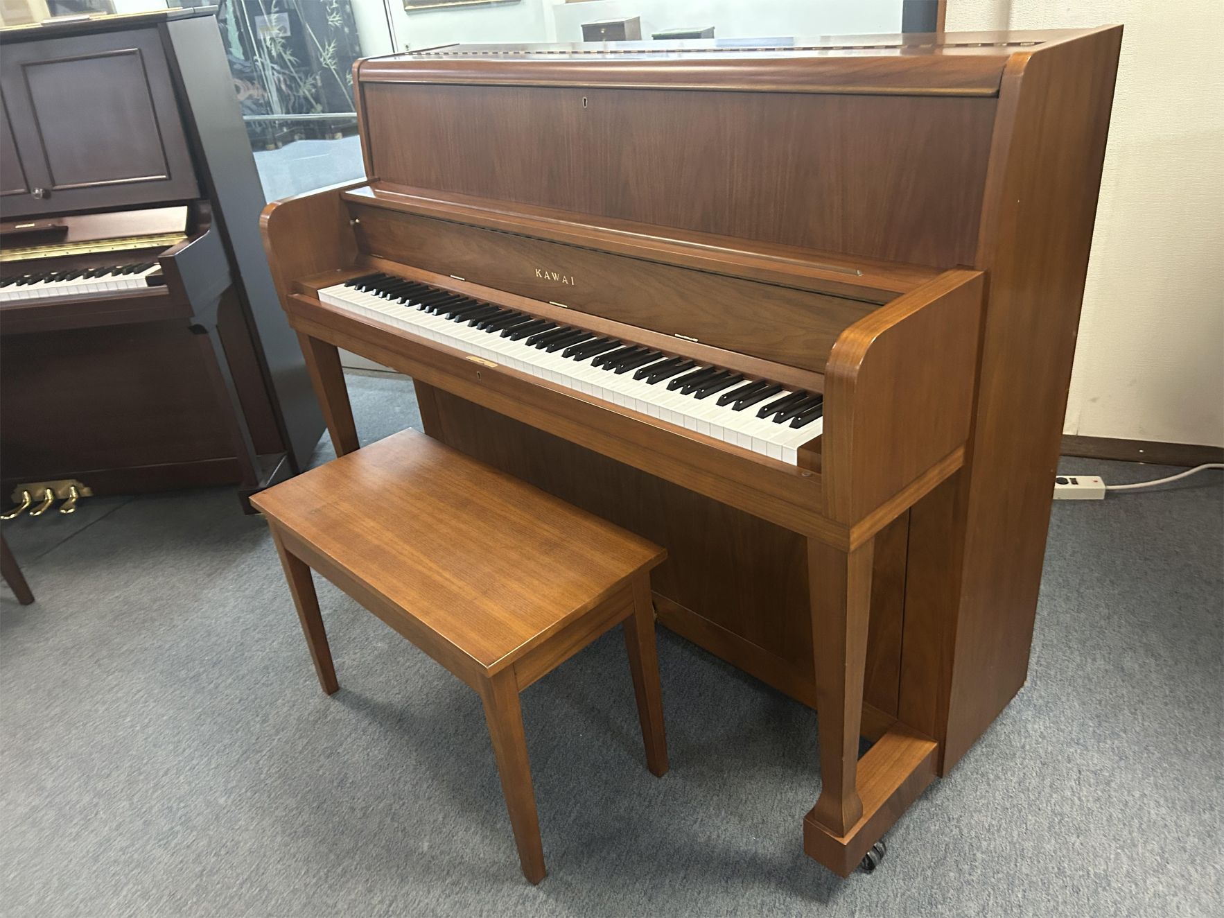 A brown upright piano with a matching bench on a gray carpet. The piano has a simple, classic design, evoking a sense of elegance and tradition.