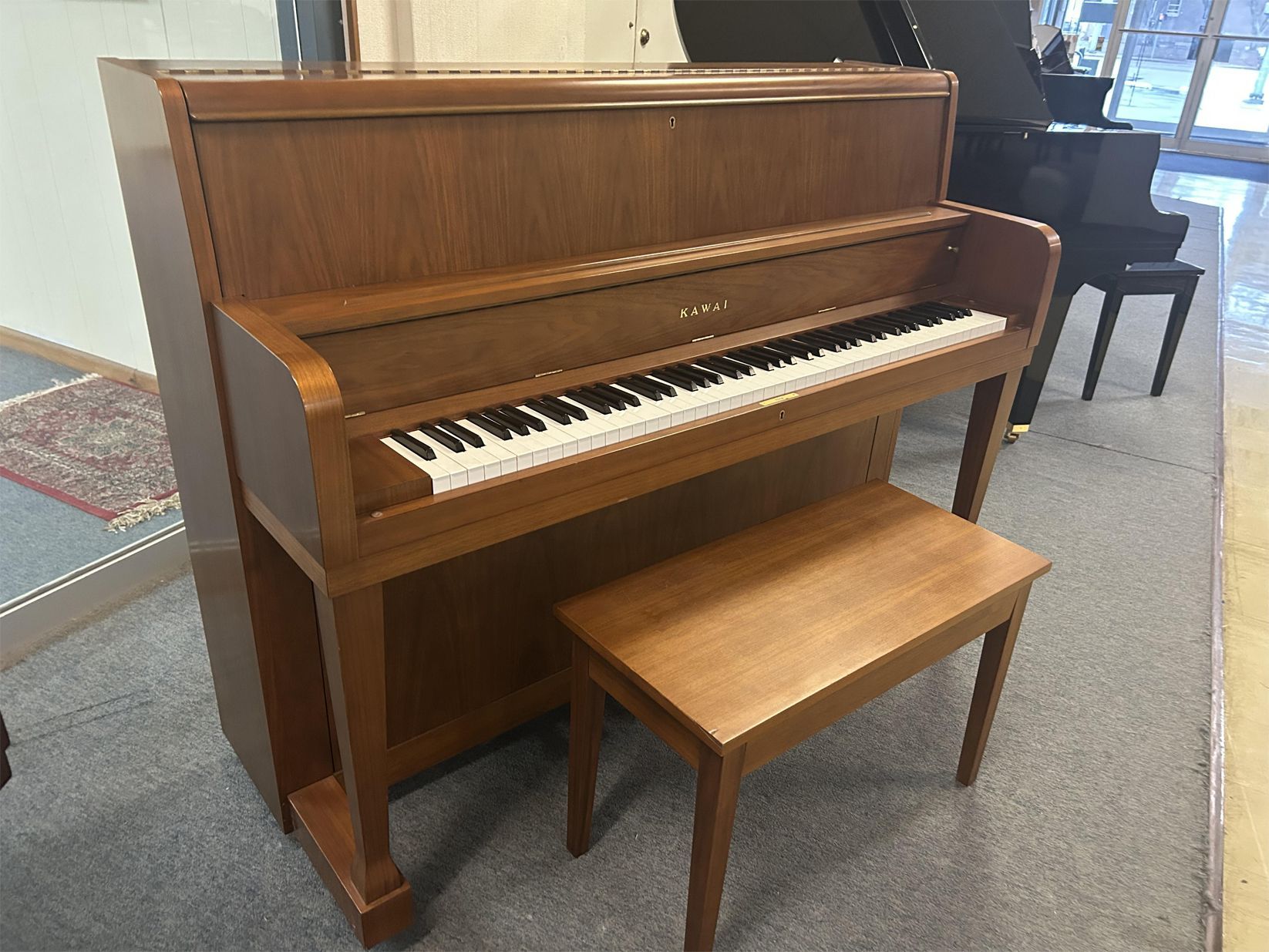 A wooden upright piano with a matching bench, featuring a warm brown finish. It is positioned on a carpet, conveying a calm and classic setting.