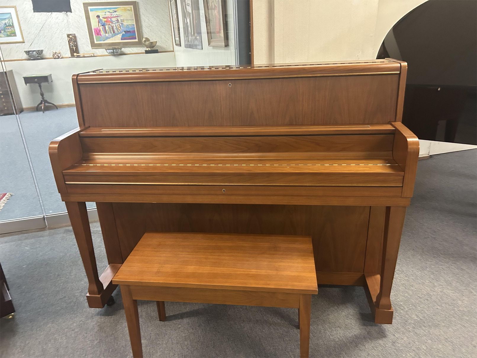 A wooden upright piano with a matching bench sits in a room with framed artwork on the walls. The setting is calm and orderly.