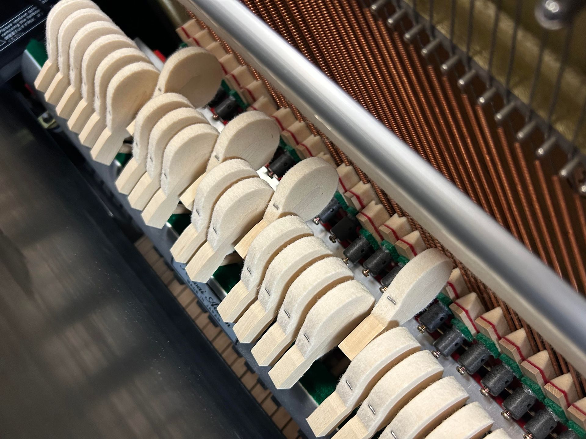 Interior view of a piano showing the hammers striking the strings. Wooden hammers are arranged in a row. Copper strings are visible.
