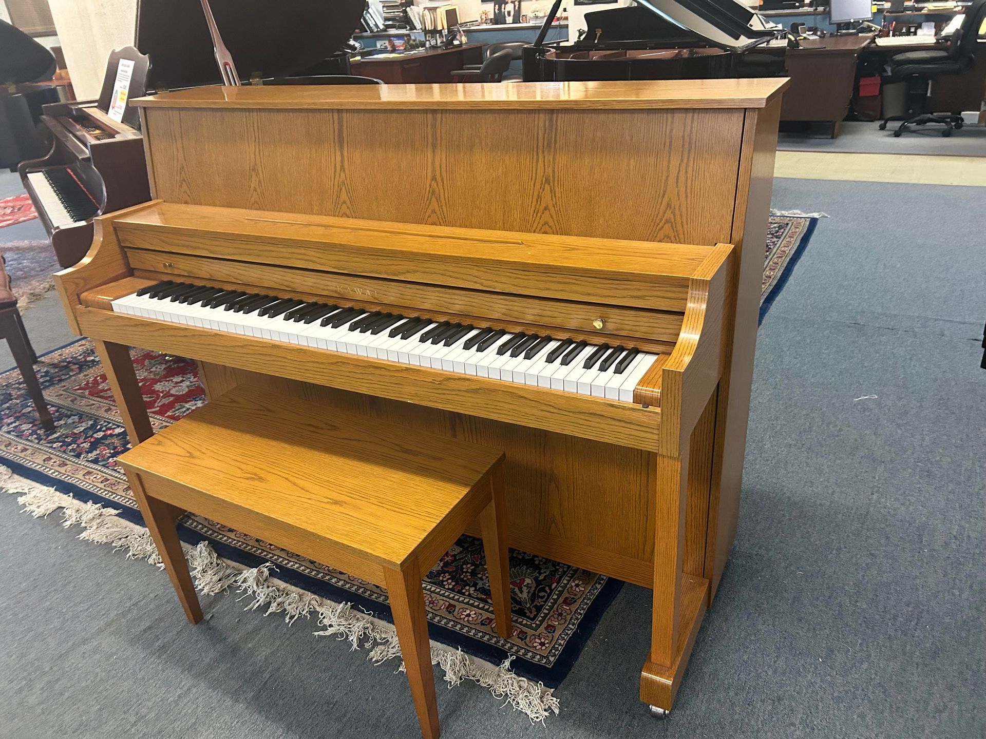 Upright piano with bench, wood grain finish, sitting on a patterned rug.
