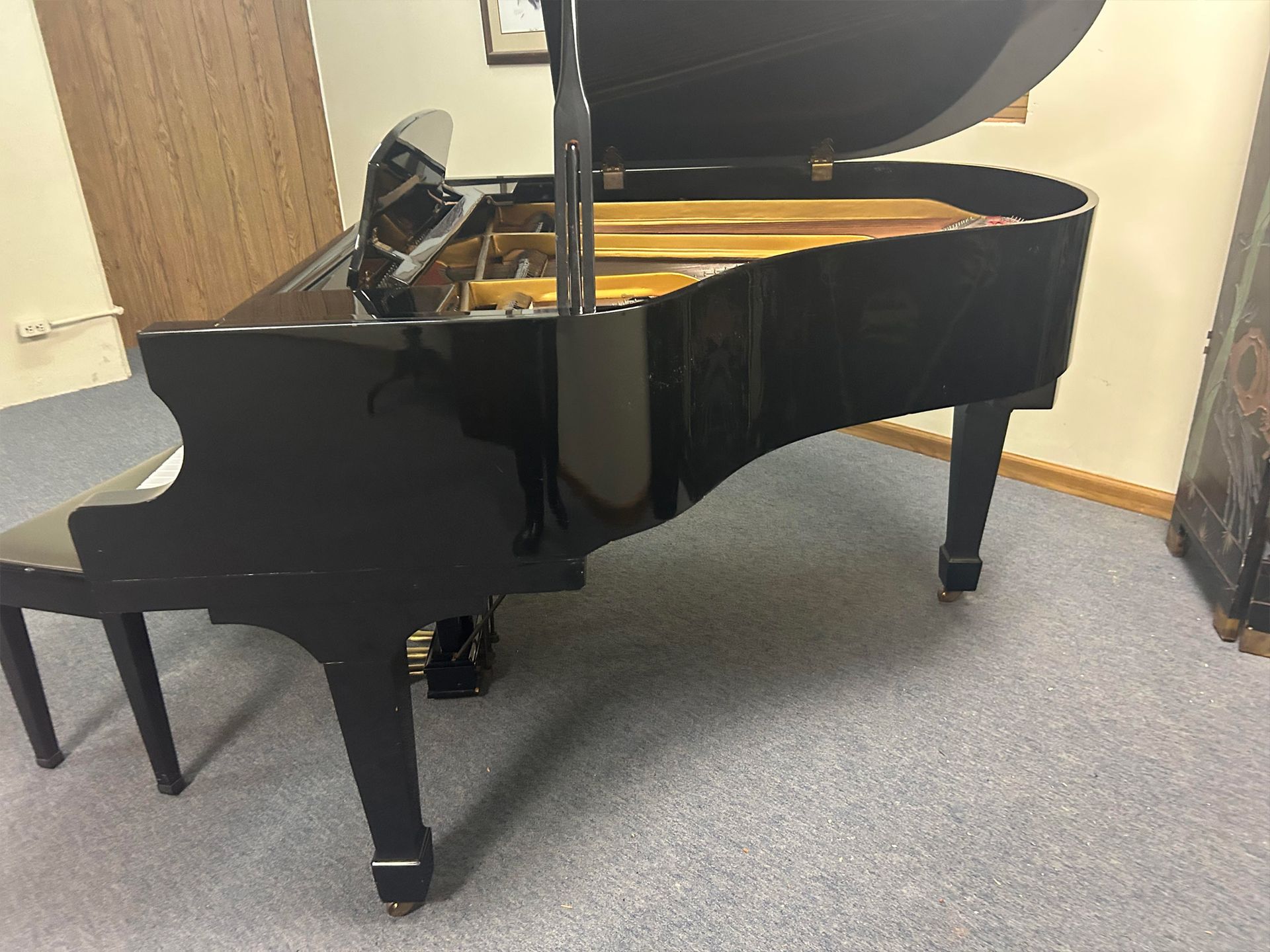 Black grand piano with open lid; black bench in a room with gray carpet.