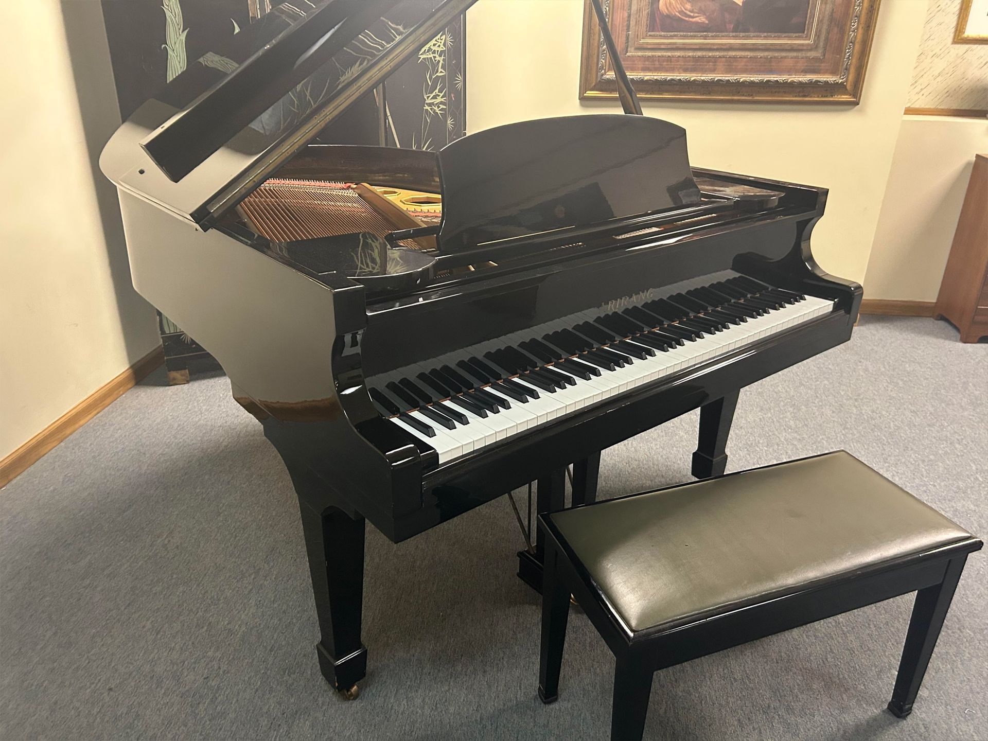 A black grand piano with the top open, a matching bench, in a room with art on the wall.