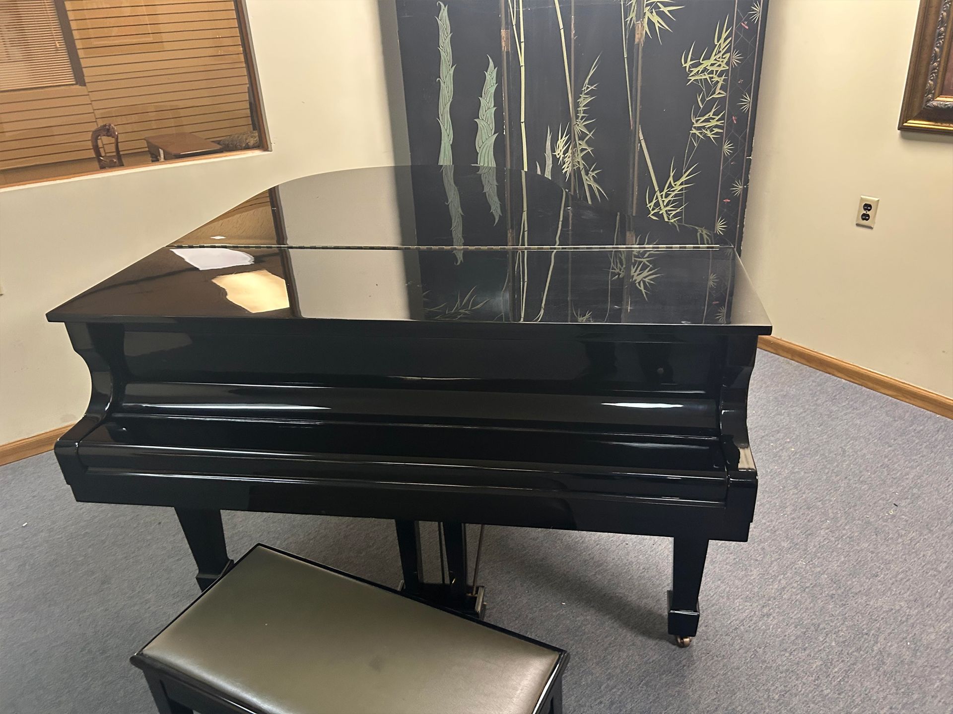 A black grand piano with a matching stool sits in a room with patterned curtains and a textured ceiling.