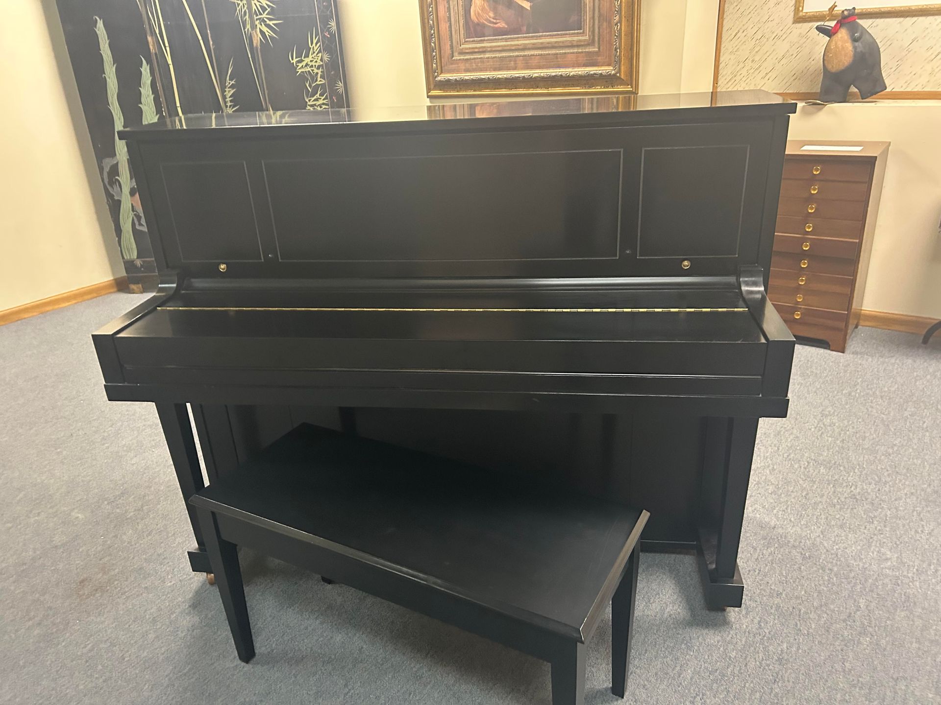 Black upright piano with matching bench. Indoors, against a wall with artwork.