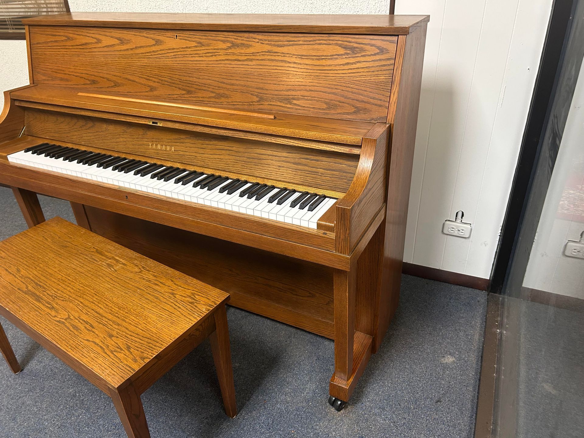 Wooden upright piano and matching bench in a room, lit by natural light.