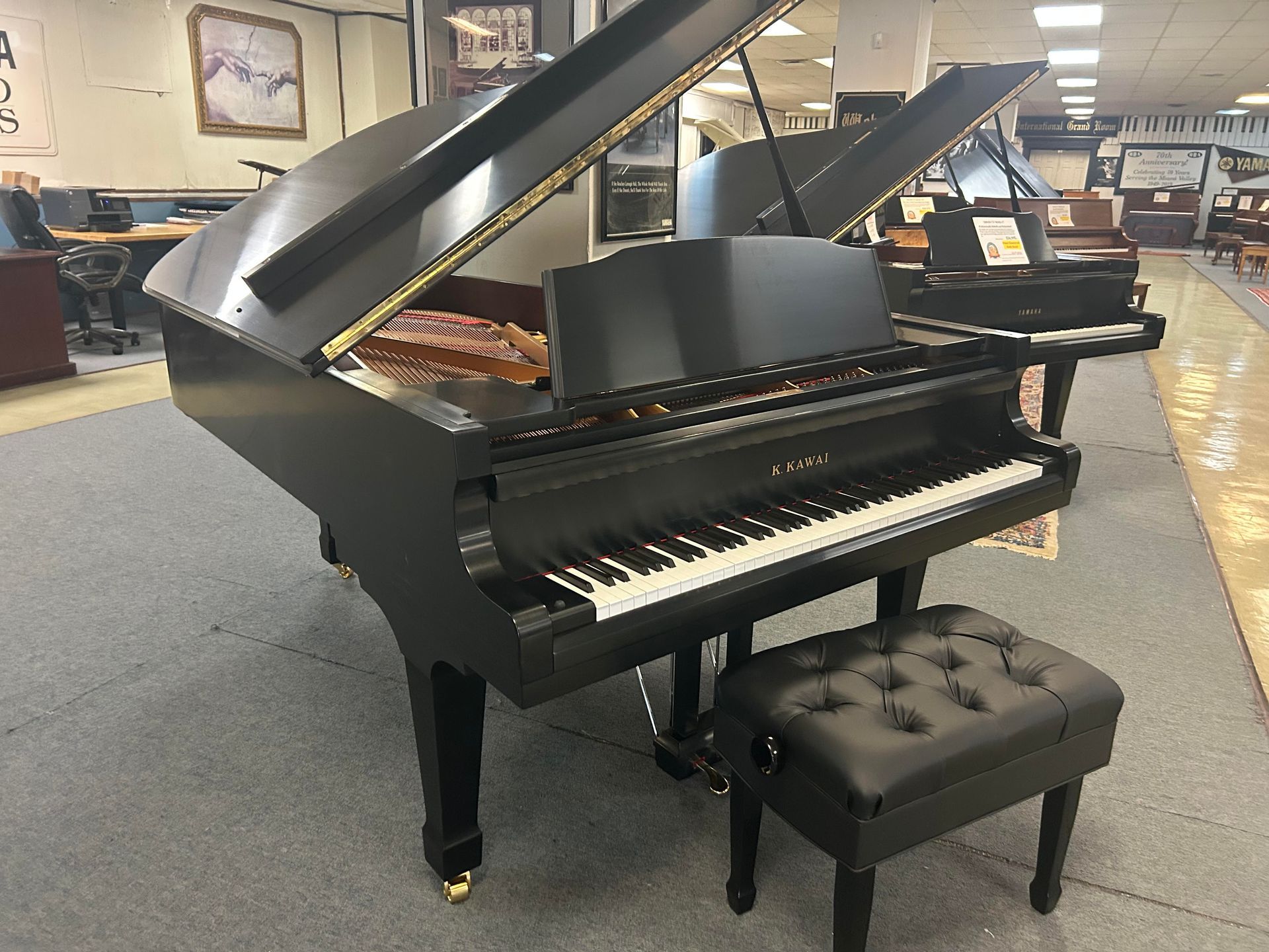 Black grand piano with open lid and a matching stool, in a showroom setting.