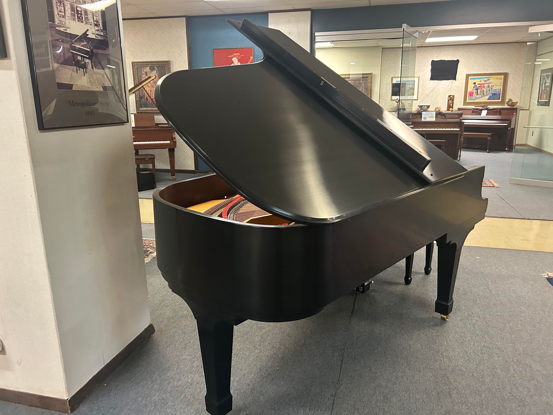 Black grand piano, lid open, in a showroom with other pianos.
