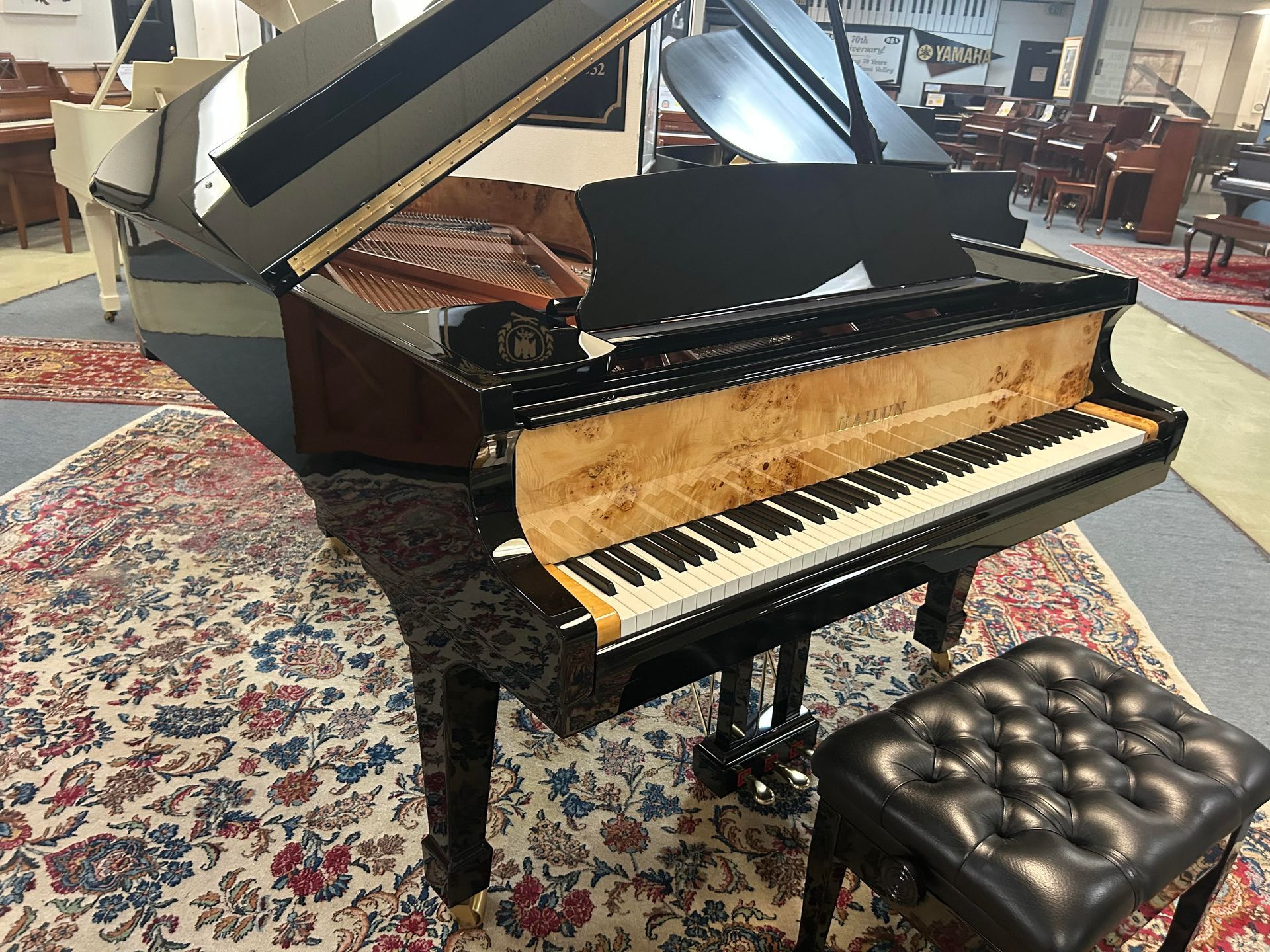 Grand piano in a showroom, black with patterned wood, keyboard exposed, and a black leather stool on a patterned rug.