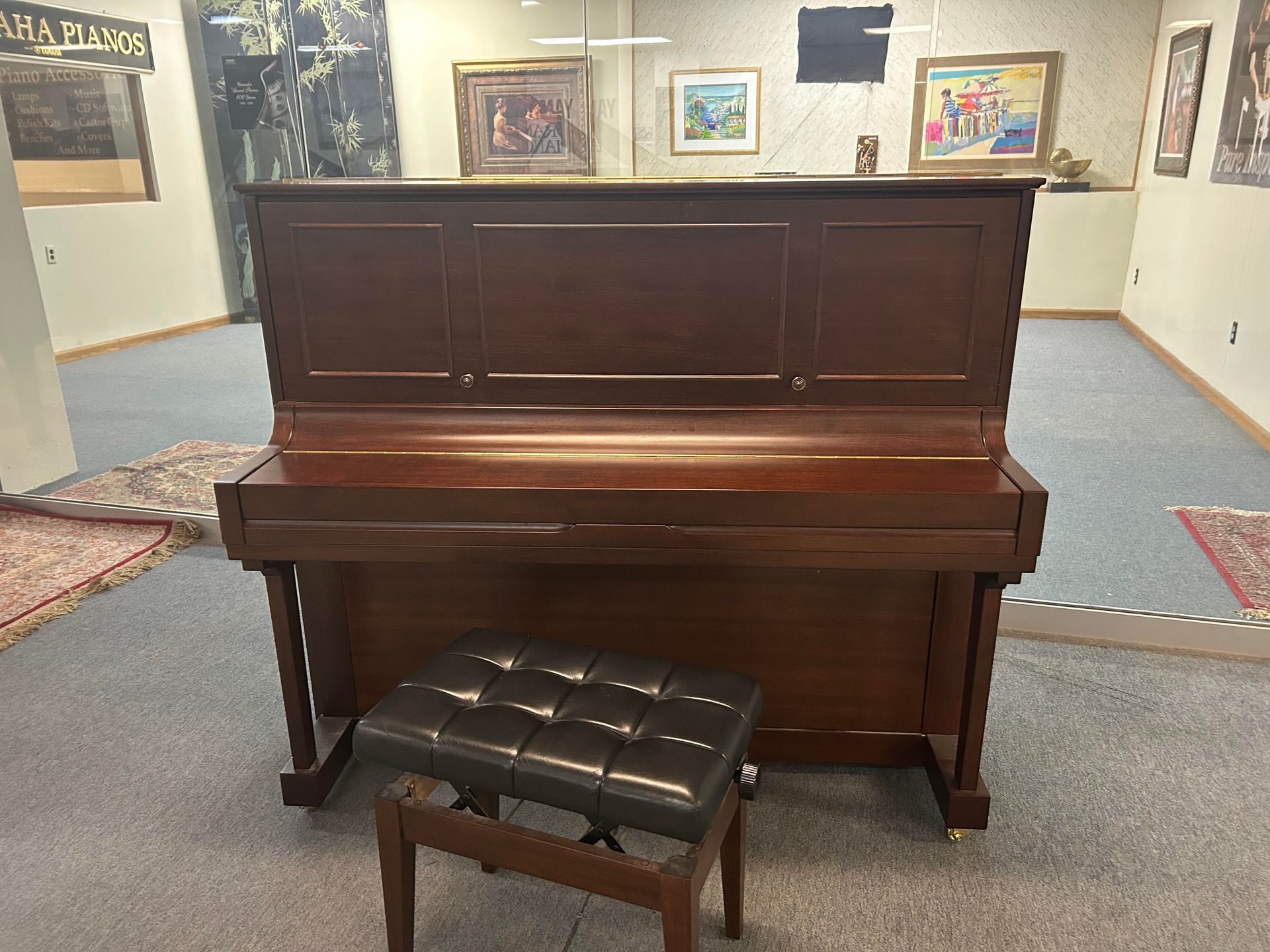 A wooden upright piano with a black leather bench in a room with artwork on the walls.