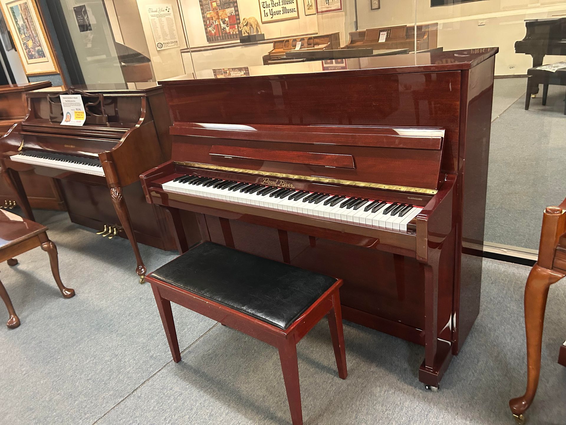 A maroon upright piano with a black bench is in a showroom. Another piano is to the left.