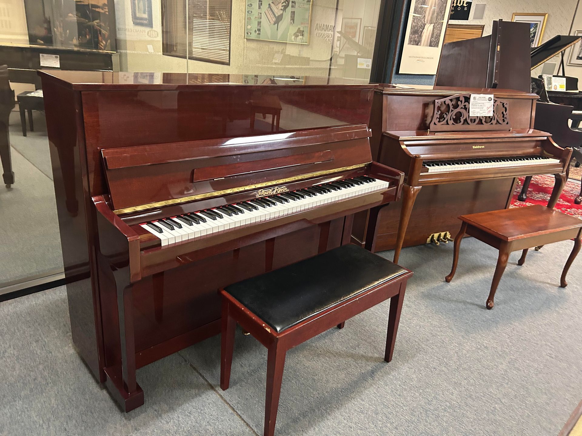 A maroon upright piano with a bench next to a smaller, antique piano and bench in a showroom.