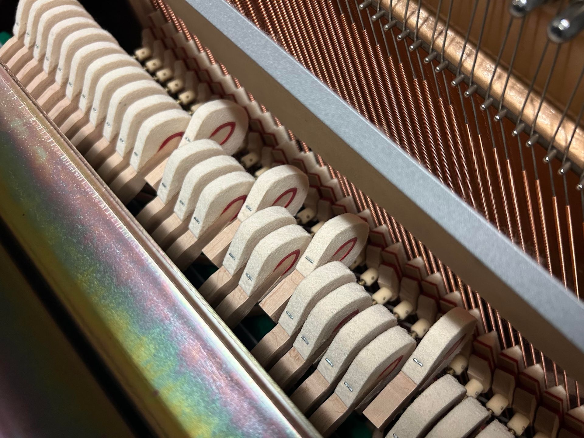Inside of a piano showing the hammers and strings. Beige felt hammers line the frame, hitting the copper strings.