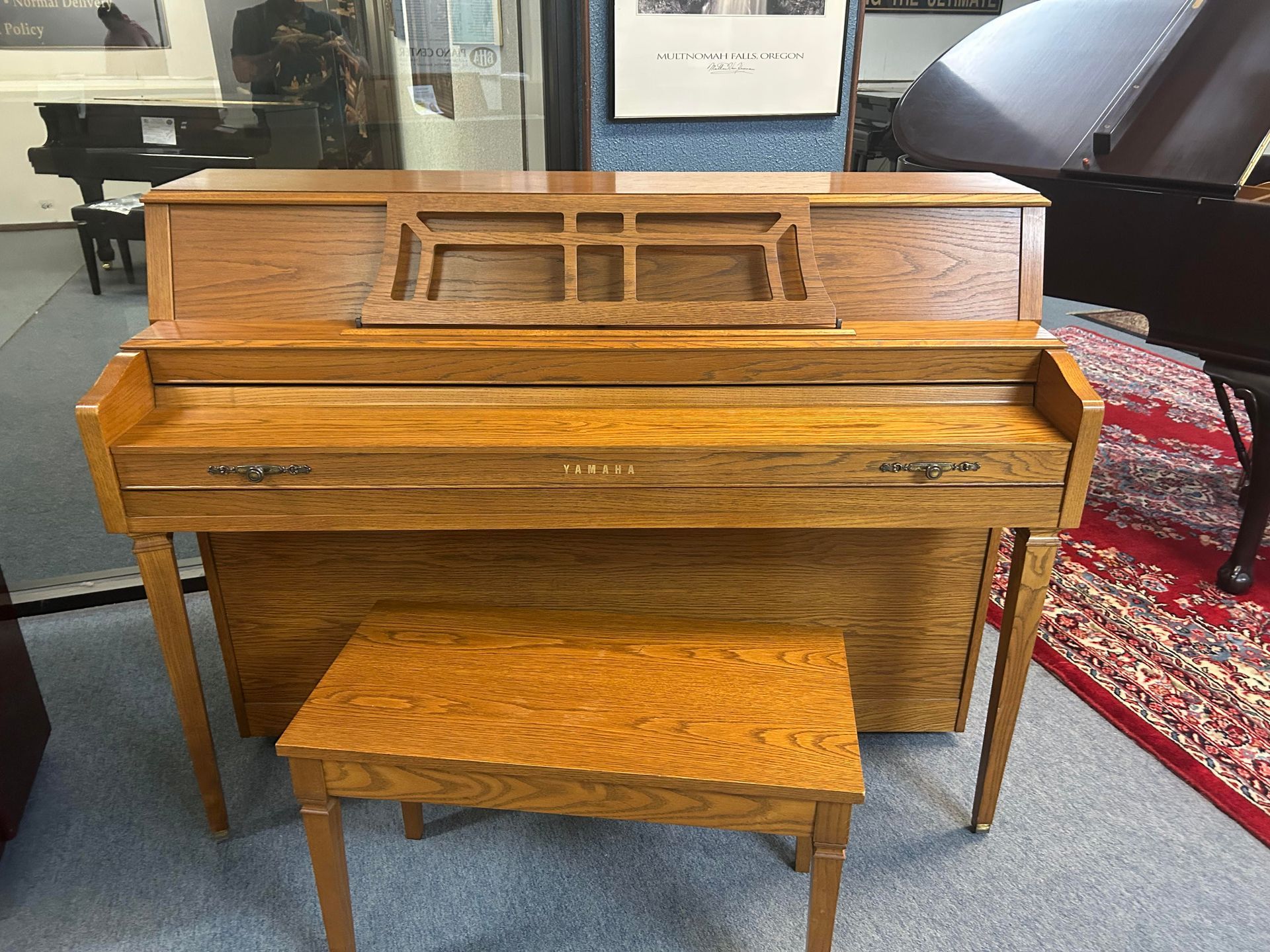 Wooden upright piano and bench in a store. The piano has a music rest and the bench is in front.