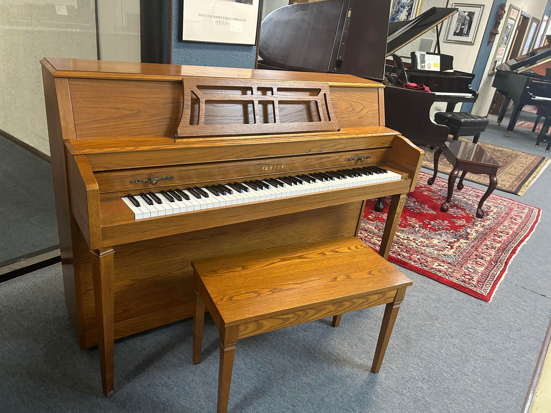 A wooden upright piano with a bench, in a store. The piano has a music rest, and sits on a patterned rug.