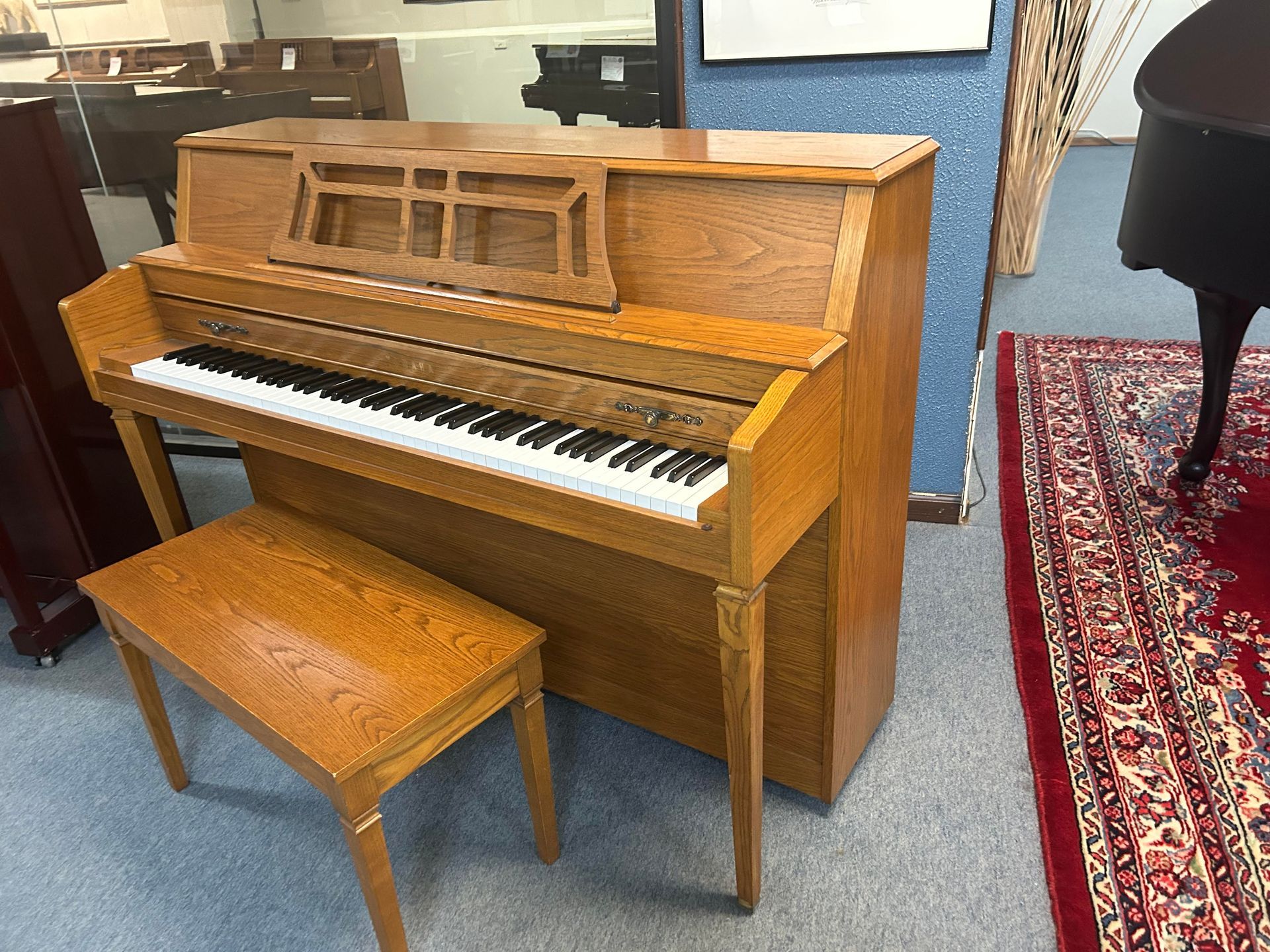 A wooden upright piano with a matching bench sits on a rug. The piano is light brown with a music stand and keyboard.