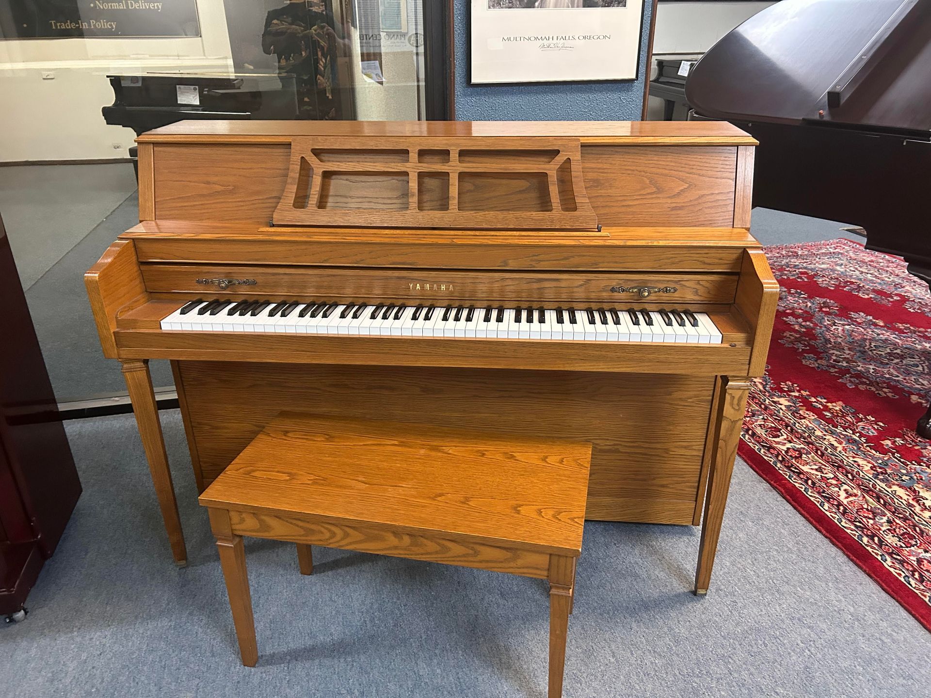 Wooden upright piano with matching bench in a room, keys visible, music stand on top.