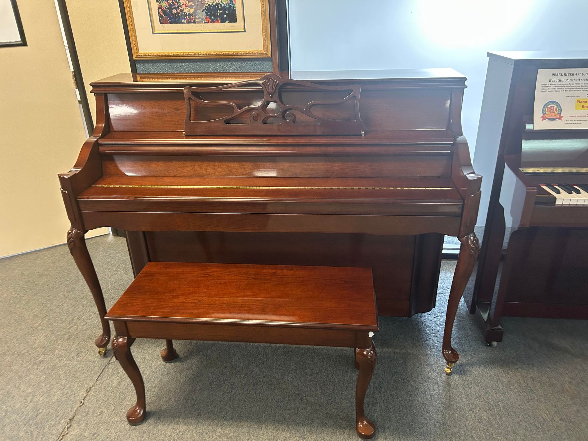 A wooden upright piano and bench. Piano is dark brown with carved details, set in a room with another piano visible.