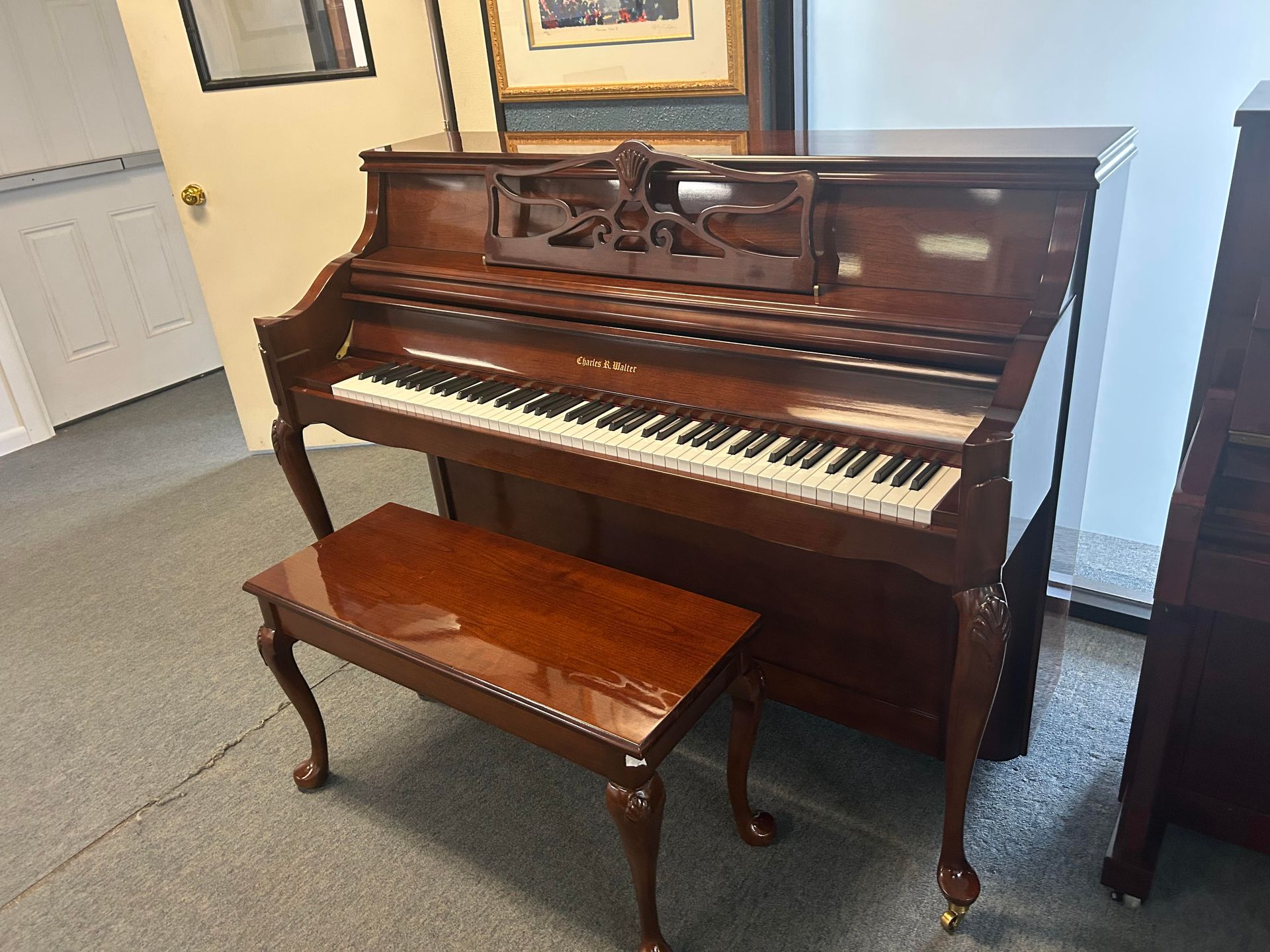 Wooden upright piano with bench; in a room with a painting on the wall.