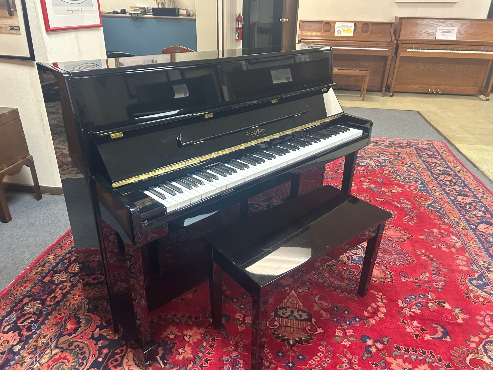 A black upright piano with a matching bench sits on a red patterned rug in a music shop.