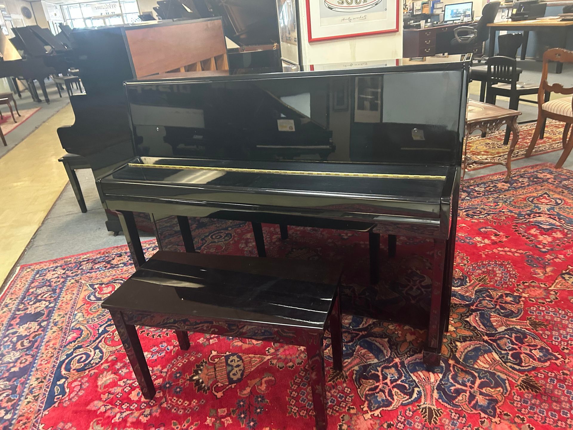 A high-gloss black upright piano and matching bench stand on a red patterned carpet in a showroom.