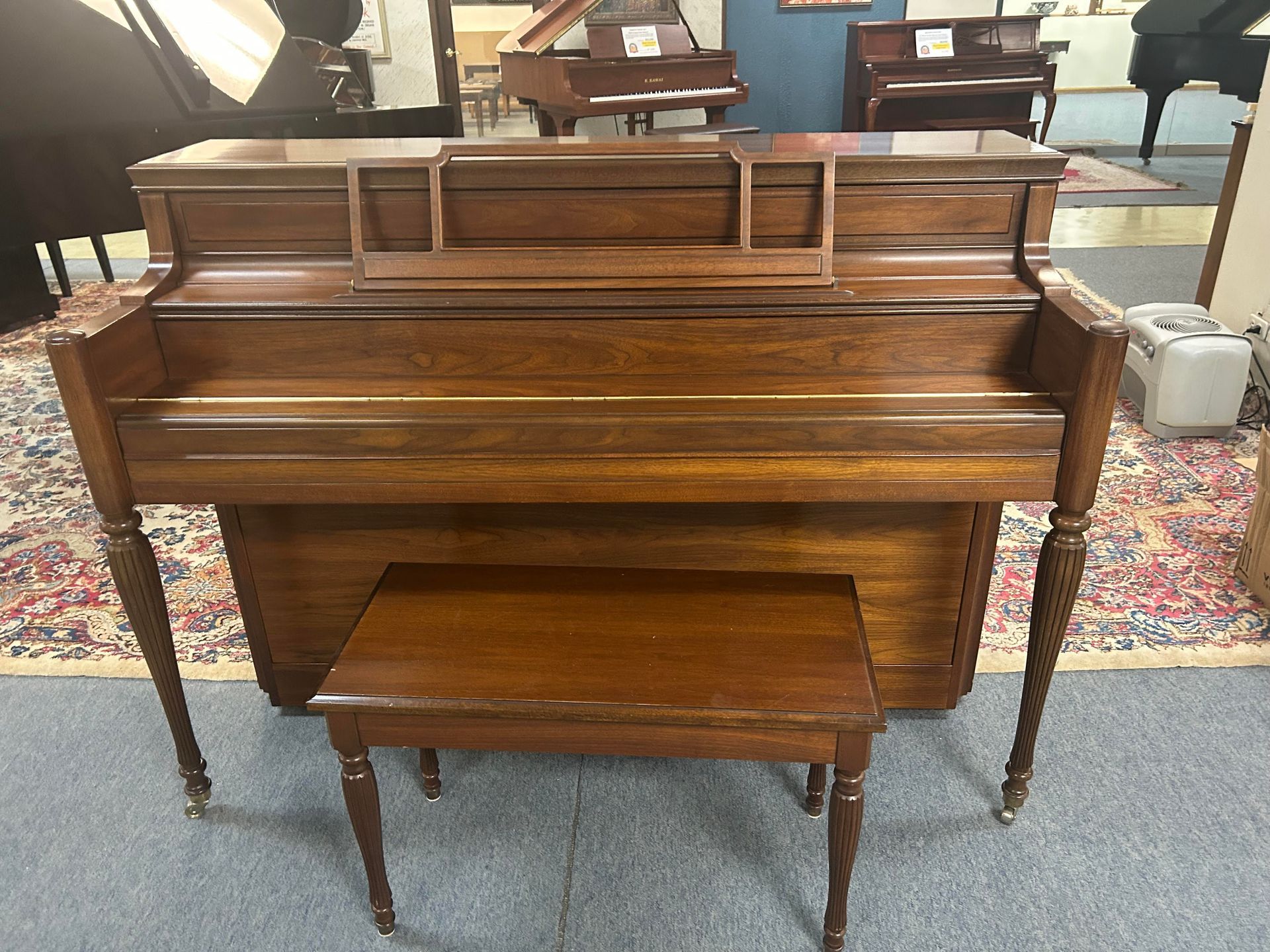 Upright wooden piano with bench, in a store setting, with a dark wood finish and ornate legs.