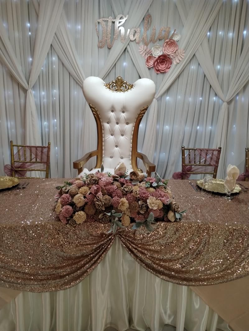 Wedding reception table with golden tablecloth, large ornate chair, floral arrangement.