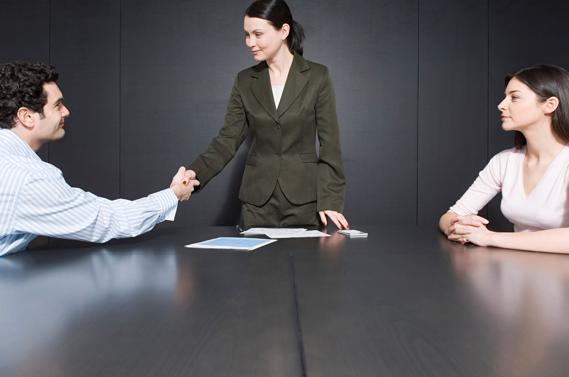 Woman shaking hands with a man over a table; woman sits nearby.