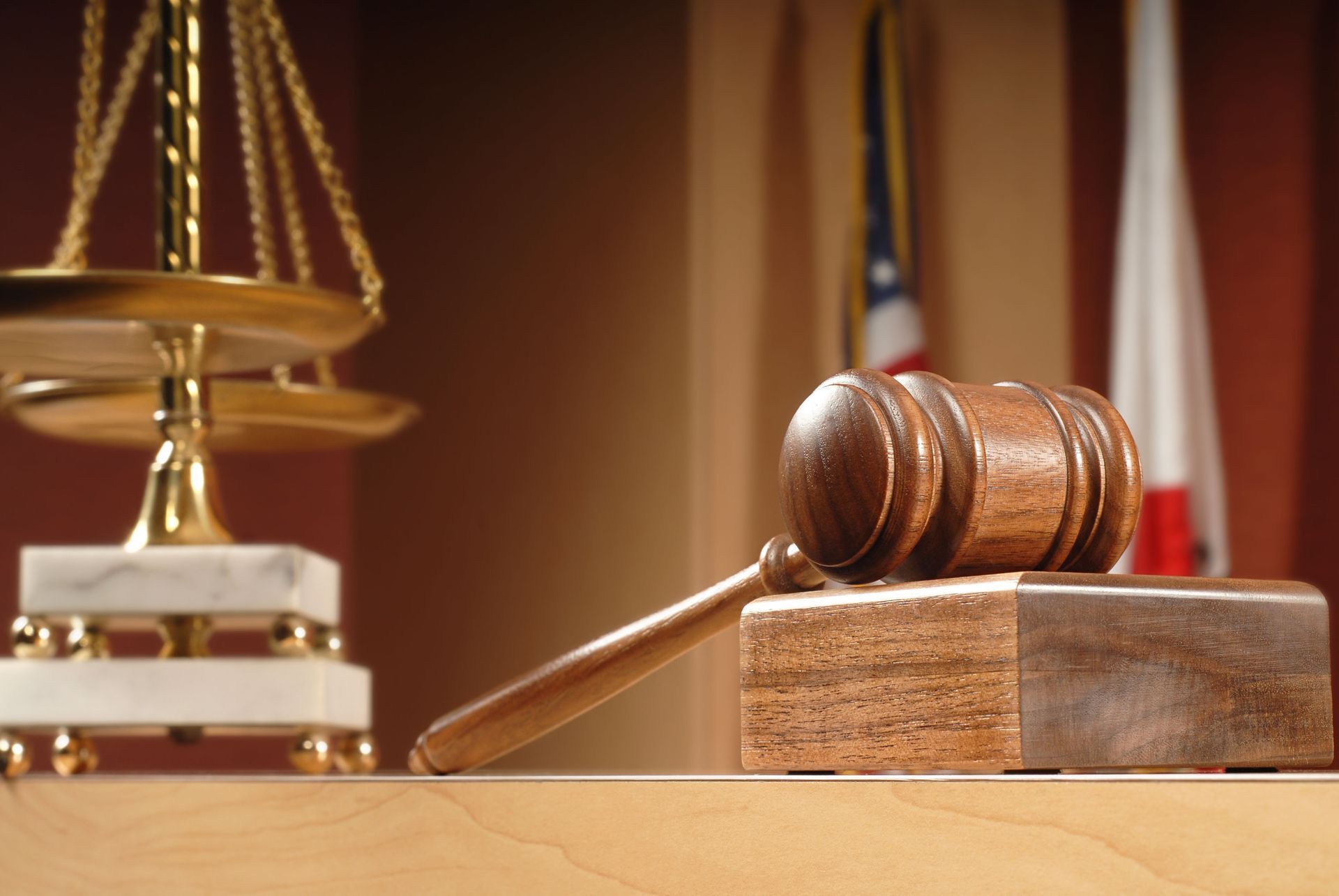 Gavel on wooden block in a courtroom setting, scales of justice and flags in background.