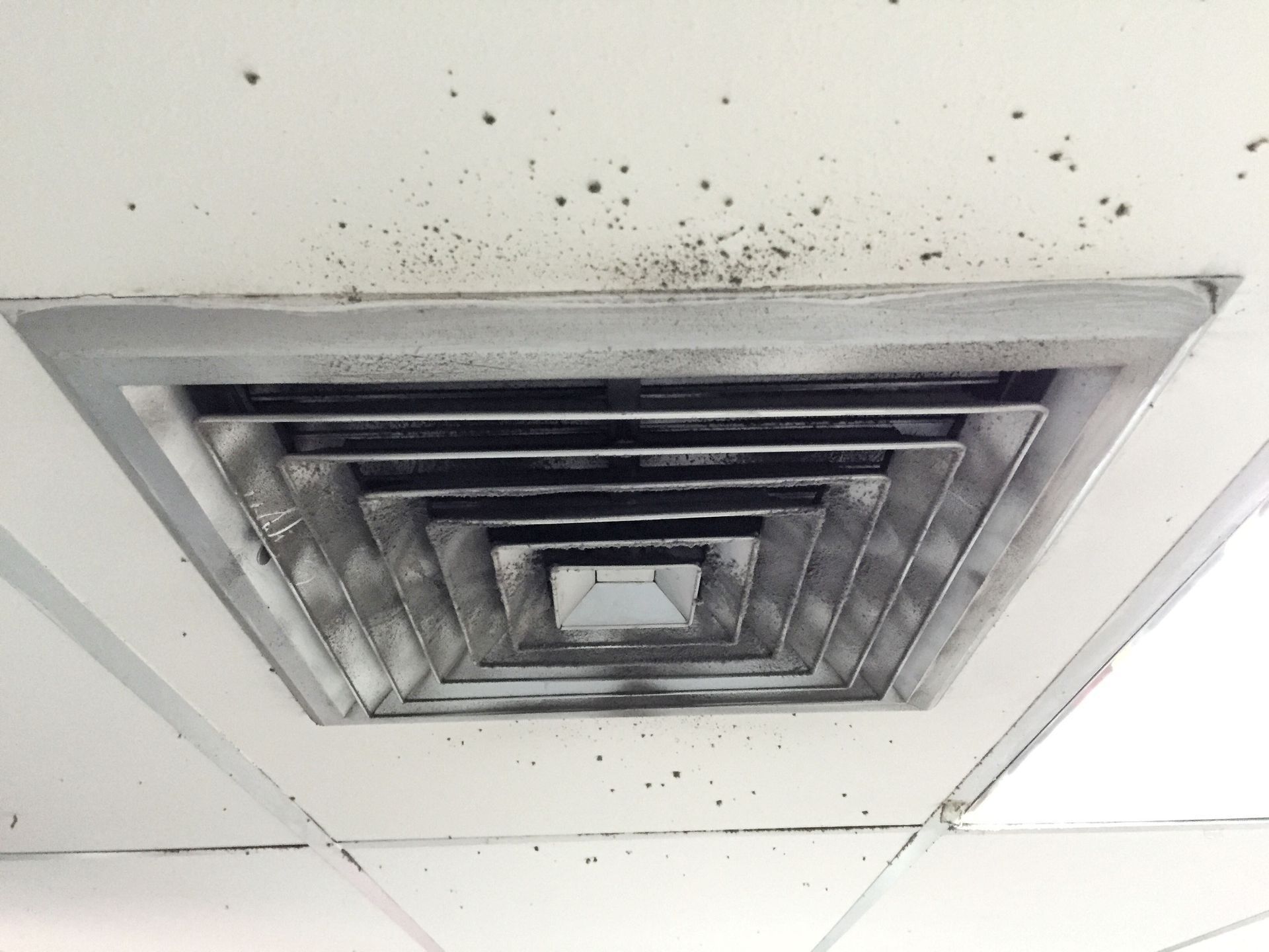 Square air vent in a white ceiling, covered in dark dust and debris.