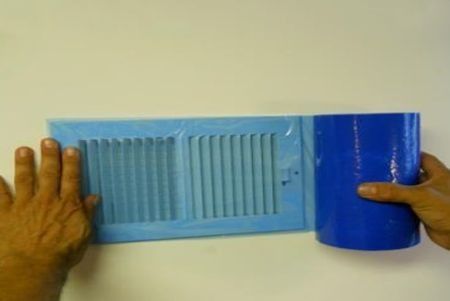 Hands holding blue vent cover and blue cylinder against a white wall.