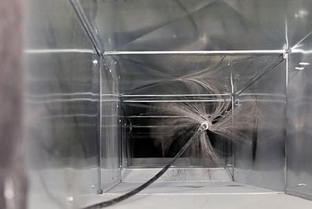 Inside of a rectangular duct, a black hose sprays dust and debris during cleaning.